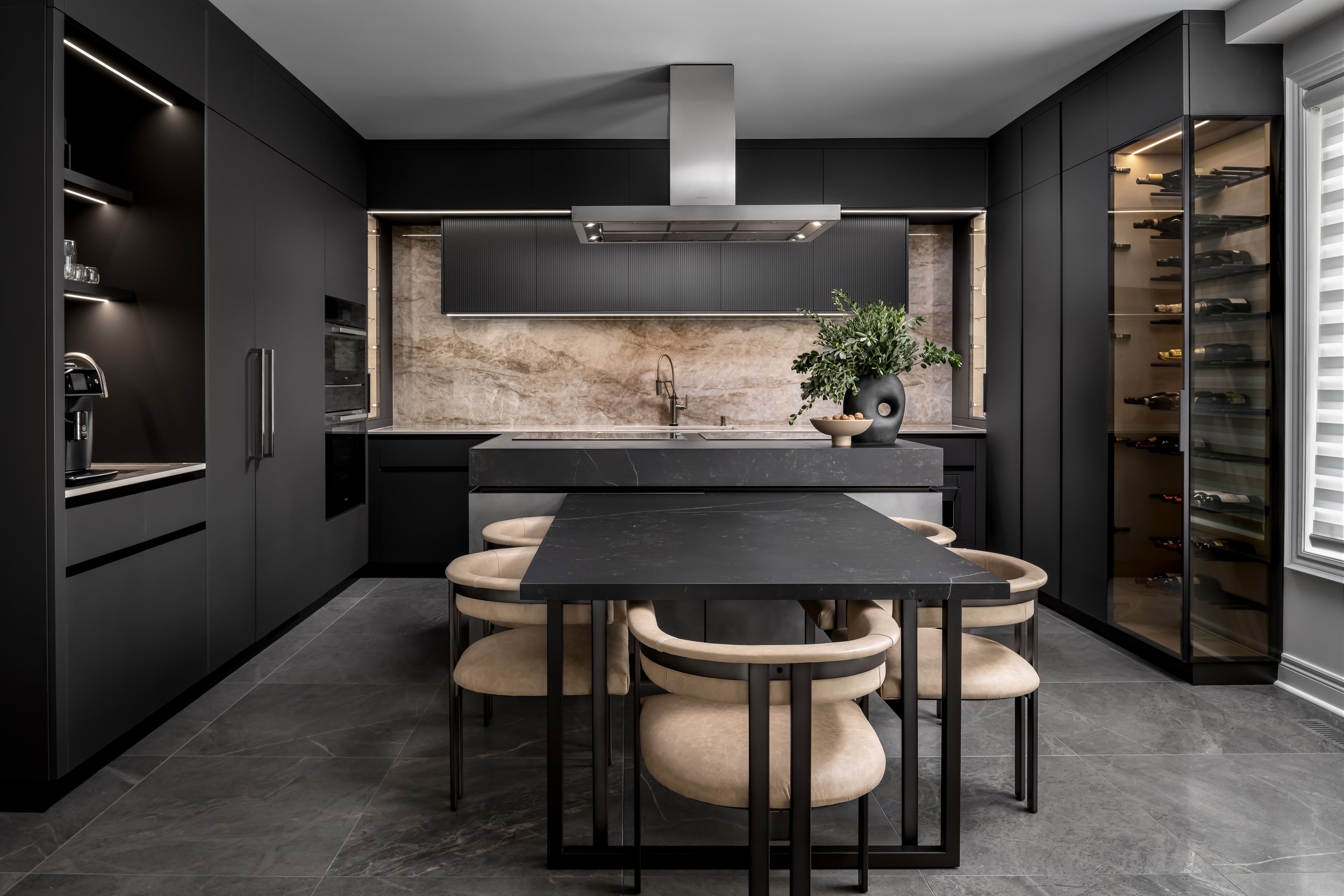 Modern kitchen with black cabinetry, marble backsplash, and a black table with beige chairs. There is a wine cooler on the right, a built-in oven, and a range hood above the stove. A decorative black vase with green plants and a small bowl are on the counter.
