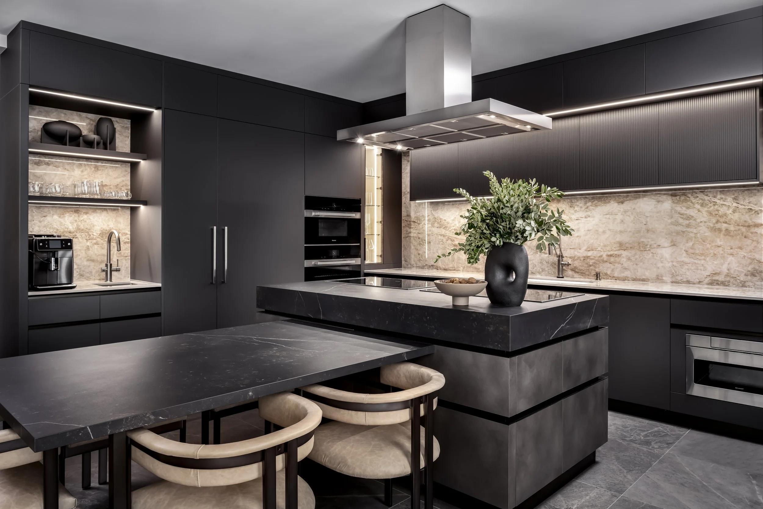 Modern kitchen with black cabinetry, a marble countertop, and a black marble island with a bowl of nuts and a vase with greenery. Stainless steel appliances and a black textured backsplash are visible.
