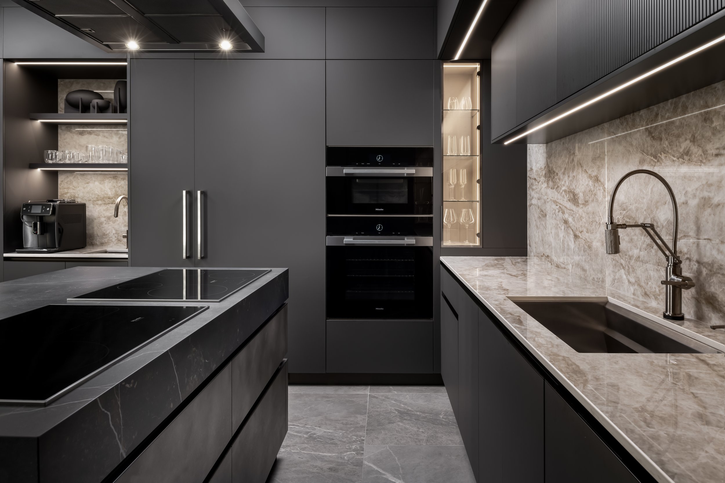 Modern kitchen with dark cabinetry, gray marble countertops, built-in oven, glass-front cabinet with glasses, and a marble backsplash.