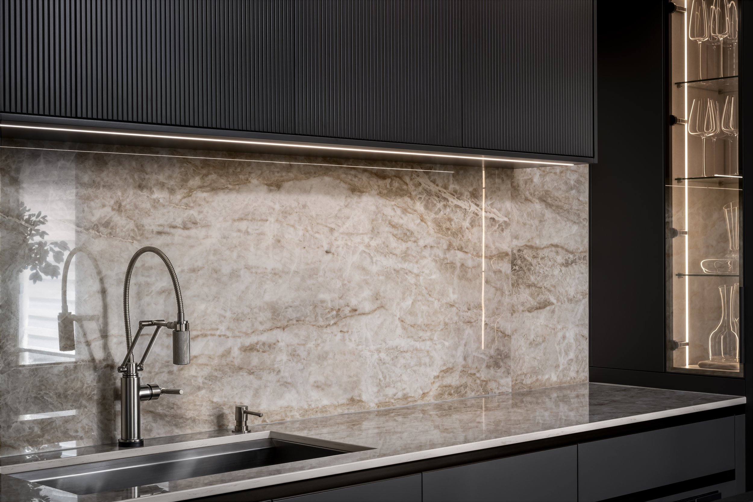 A modern kitchen with black cabinets, a marble backsplash, a stainless steel sink with a flexible faucet, and a glass-fronted cabinet with wine glasses inside, accented with soft lighting.