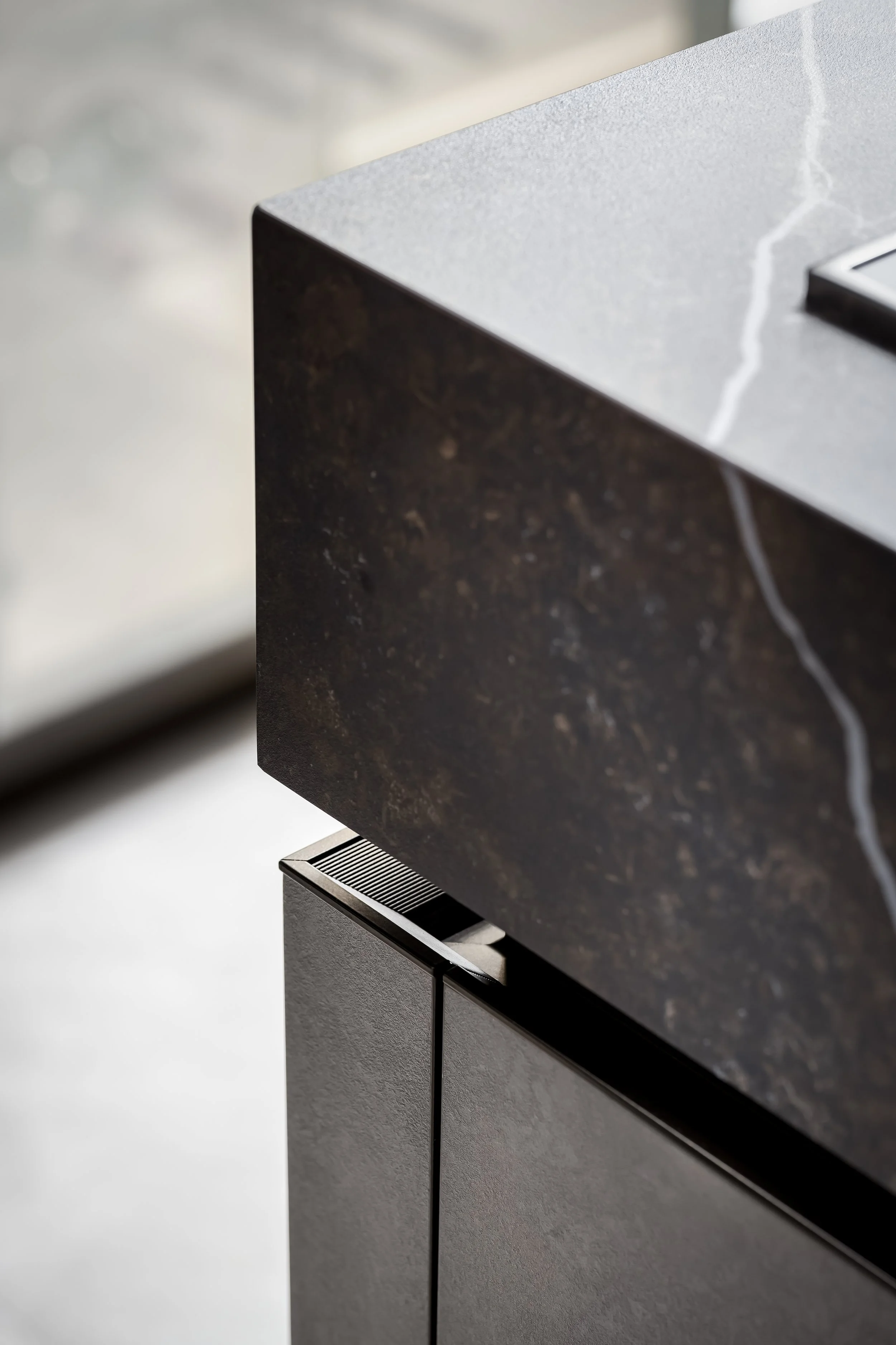 Close-up of a dark kitchen countertop with a textured surface, a section of a black and gray cabinet below, and a glimpse of a metallic kitchen appliance at the bottom. The background appears to be a light-colored wall or surface.