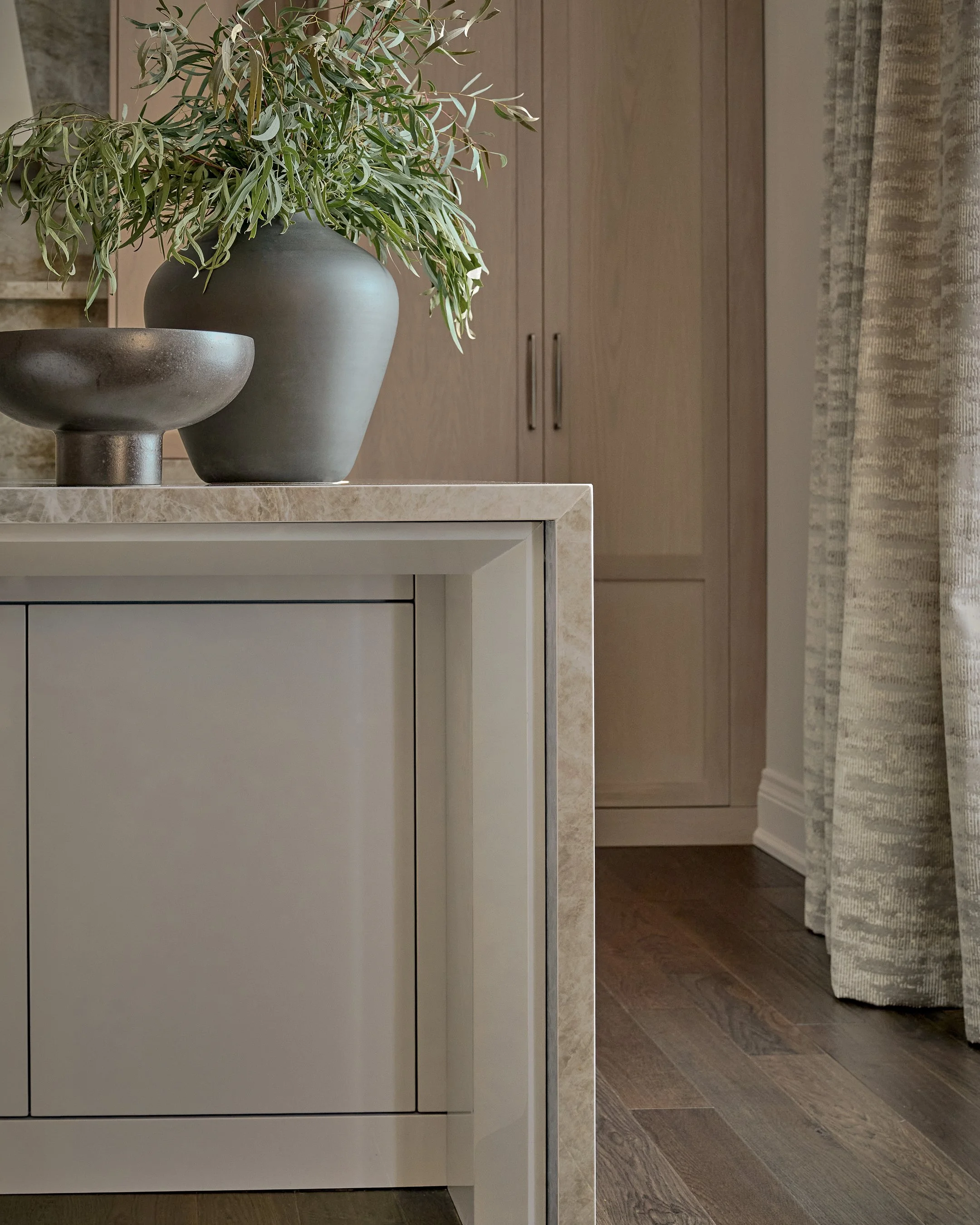 A white cabinet with a marble top features two decorative vases, one with a matte dark finish and the other with a textured metallic surface, holding greenery. In the background, there are wooden cabinets and a patterned curtain on the right side.