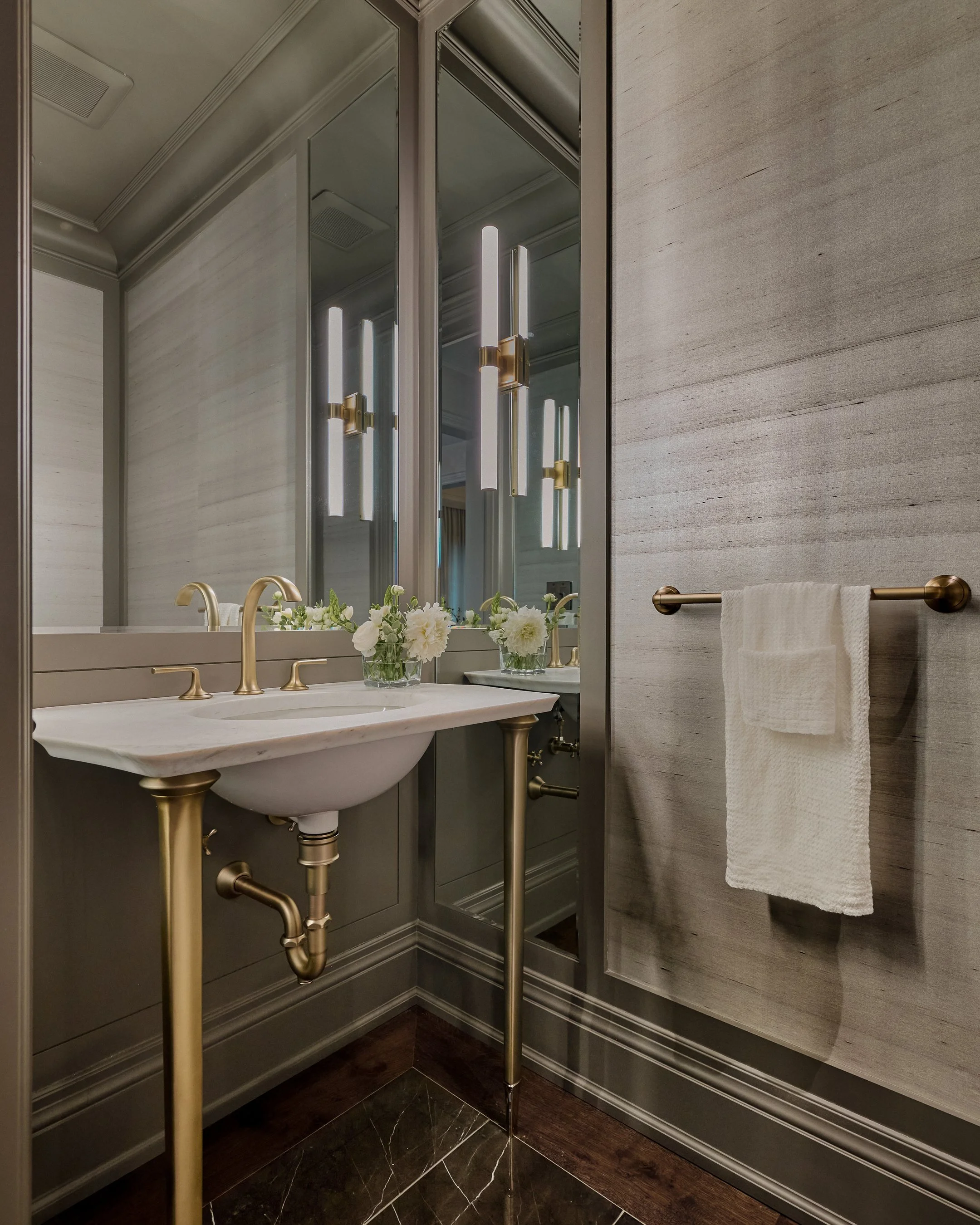 Modern bathroom vanity area with gold fixtures, a marble countertop, floral arrangements, a large mirror, and a textured wall with a towel rack.