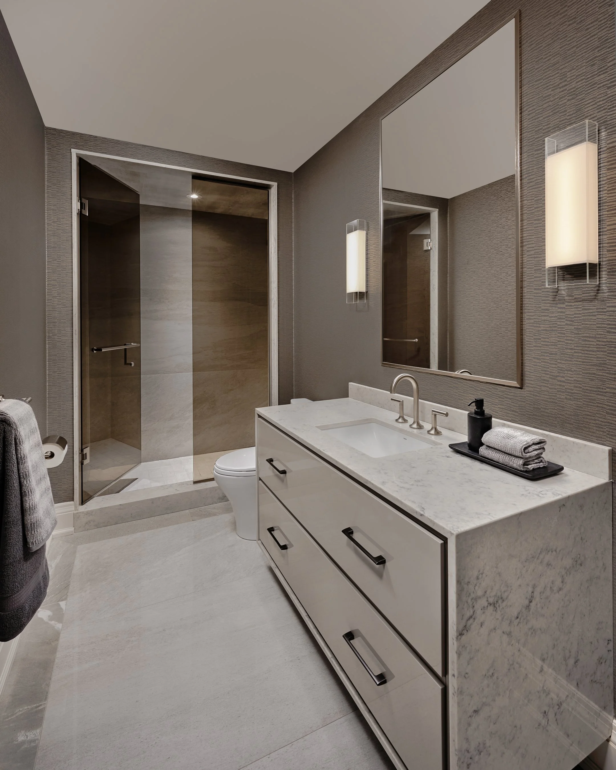 Modern bathroom with a sink vanity, large mirror, wall sconce, toilet, and walk-in shower with glass doors, gray textured walls, and light-colored floor tiles.