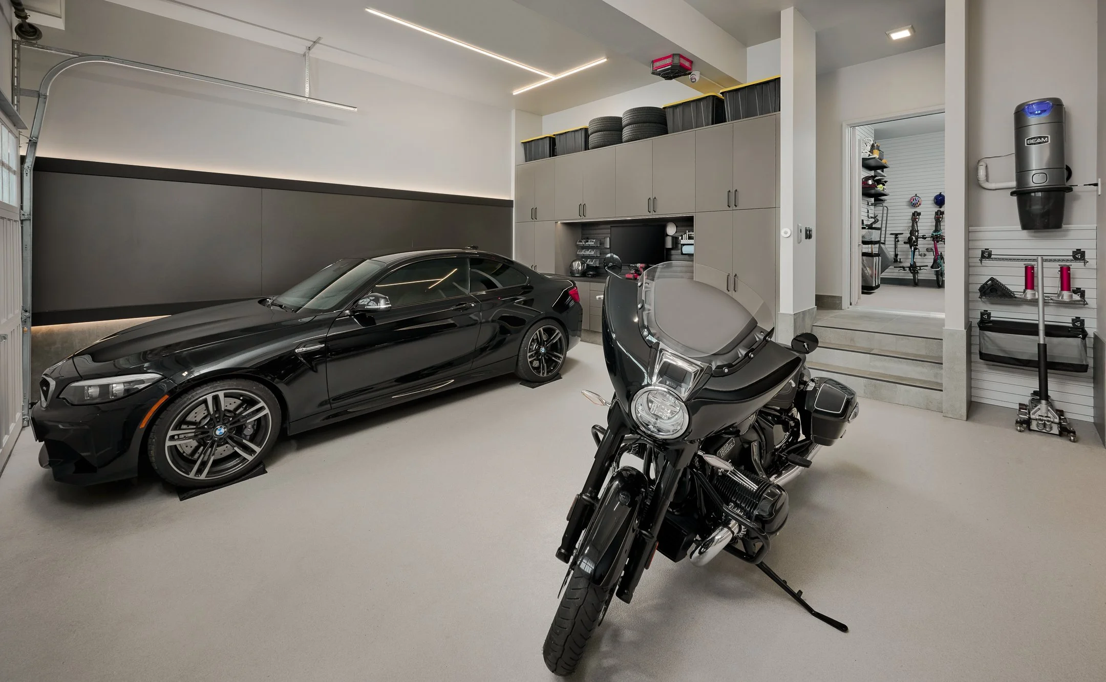 A modern garage with a black sports car and a black motorcycle parked inside, with storage cabinets, tires, and tools on the walls.