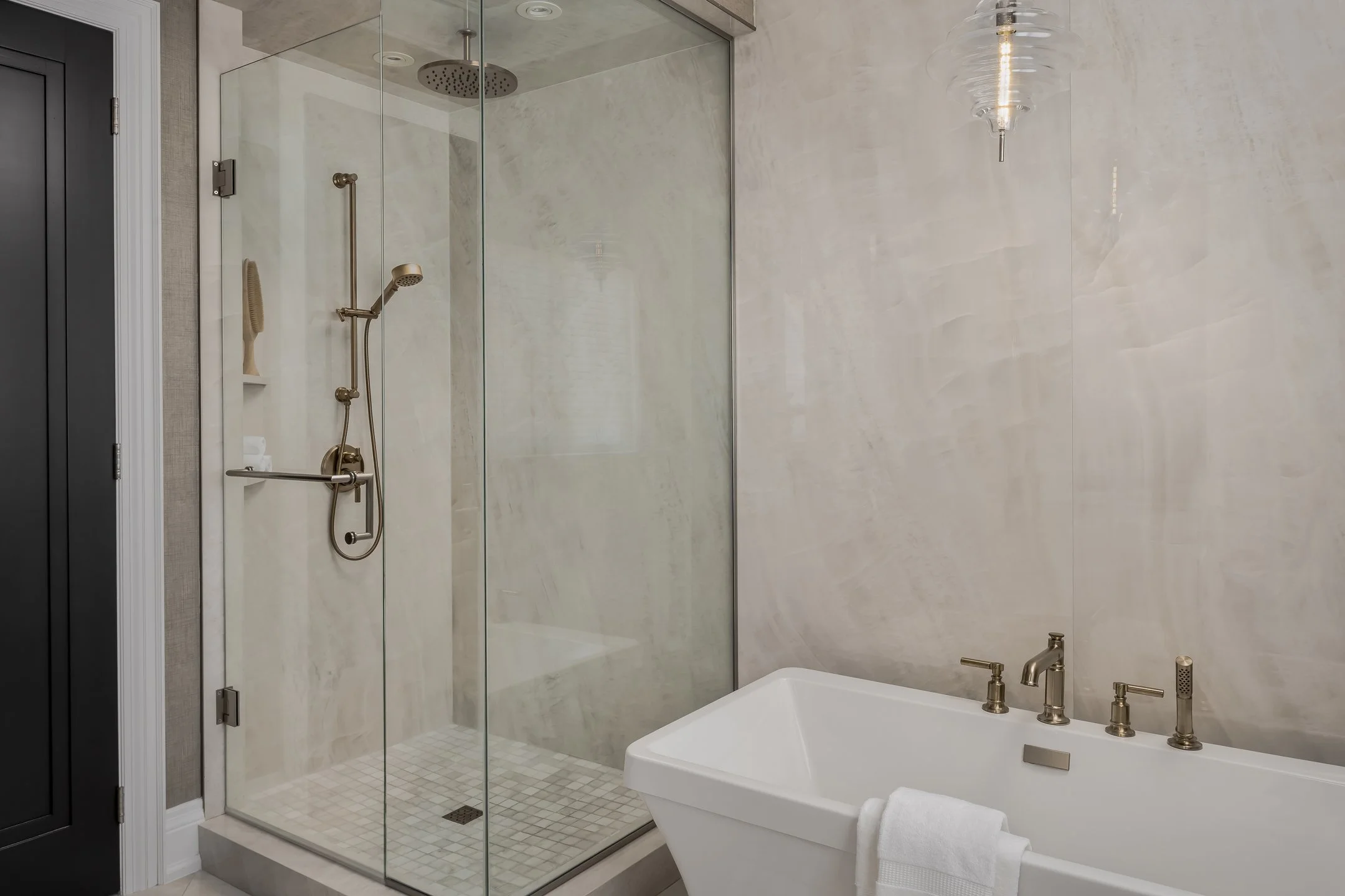 Modern bathroom with a glass-enclosed shower area that has metallic fixtures, a shower head, and a handheld shower wand. Adjacent to the shower is a white bathtub with metallic fixtures and a white towel draped over its side. The wall is finished with light-colored marble or tile, and there is a decorative glass pendant light hanging from the ceiling.