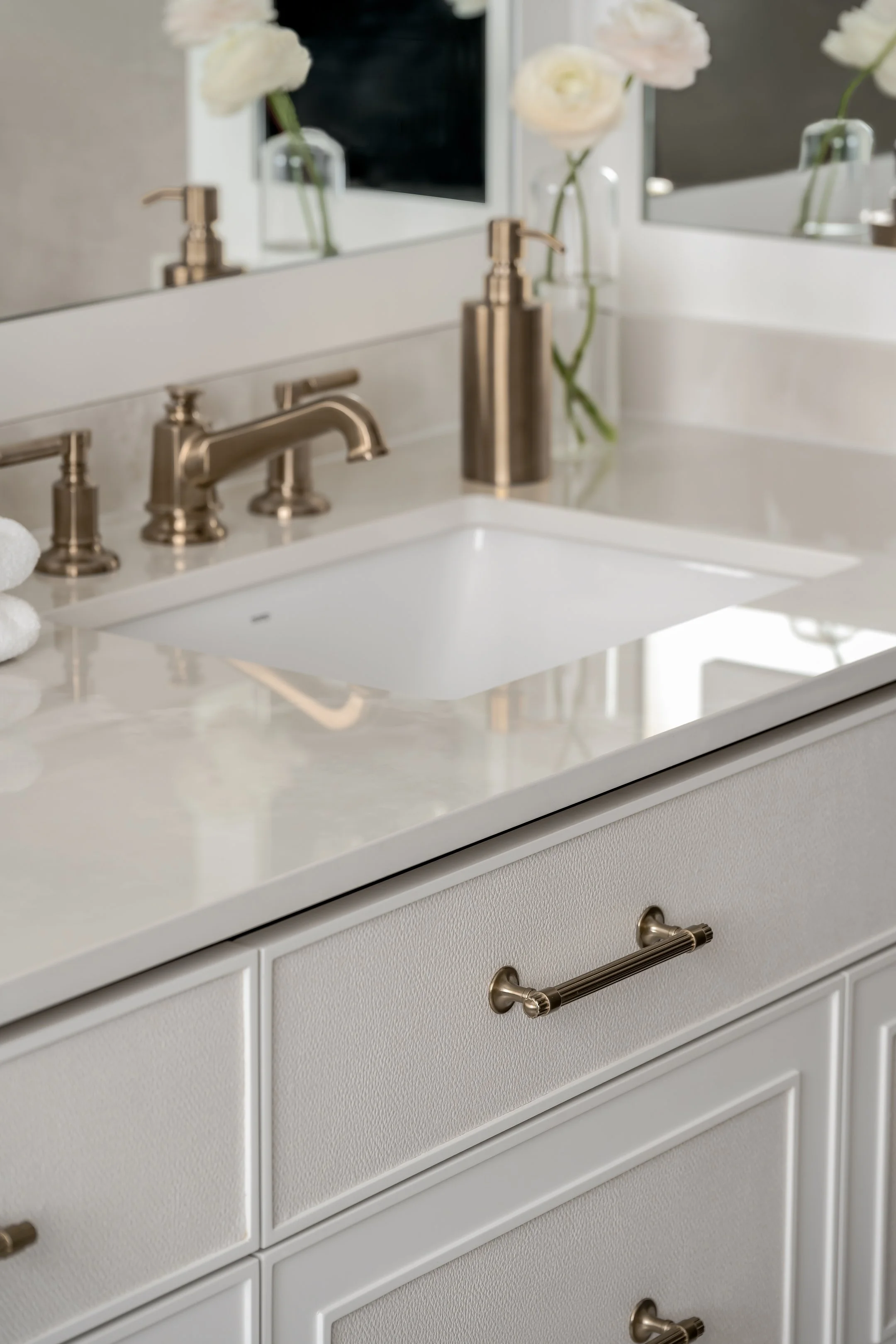 Close-up of a white bathroom vanity with a rectangular sink, brass faucet, and two cabinet drawers, with a mirror above reflecting white flowers in a vase.