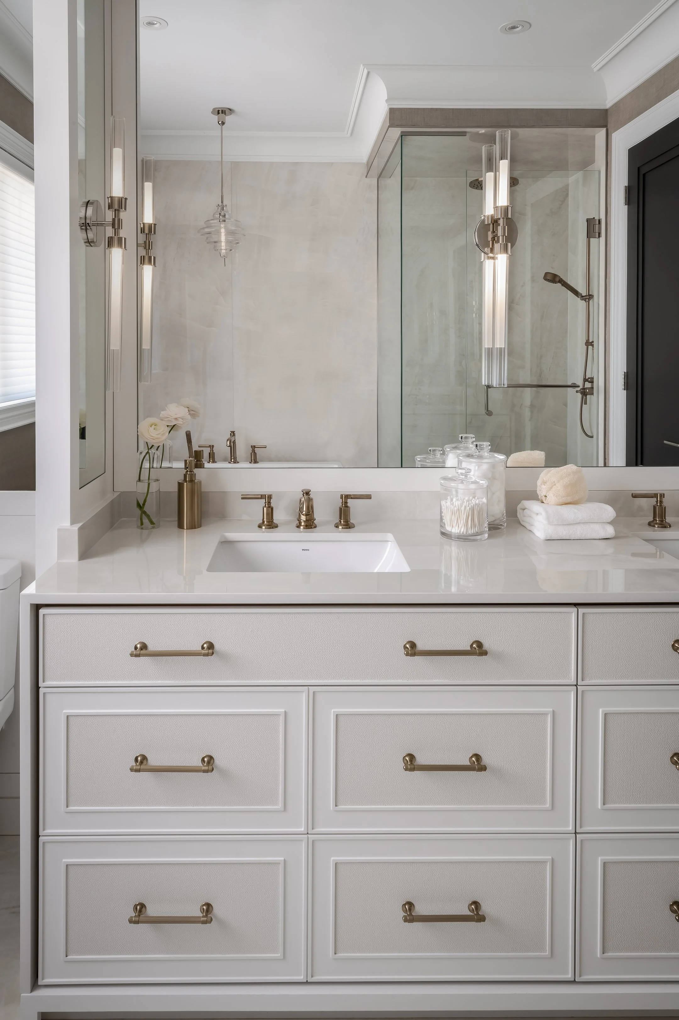 A bathroom vanity with a white countertop, brass hardware, and a mirror. On the countertop, there are glass jars with cotton swabs and balls, a rolled towel, and a vase with white flowers. The background shows a walk-in shower with glass doors and modern lighting fixtures.