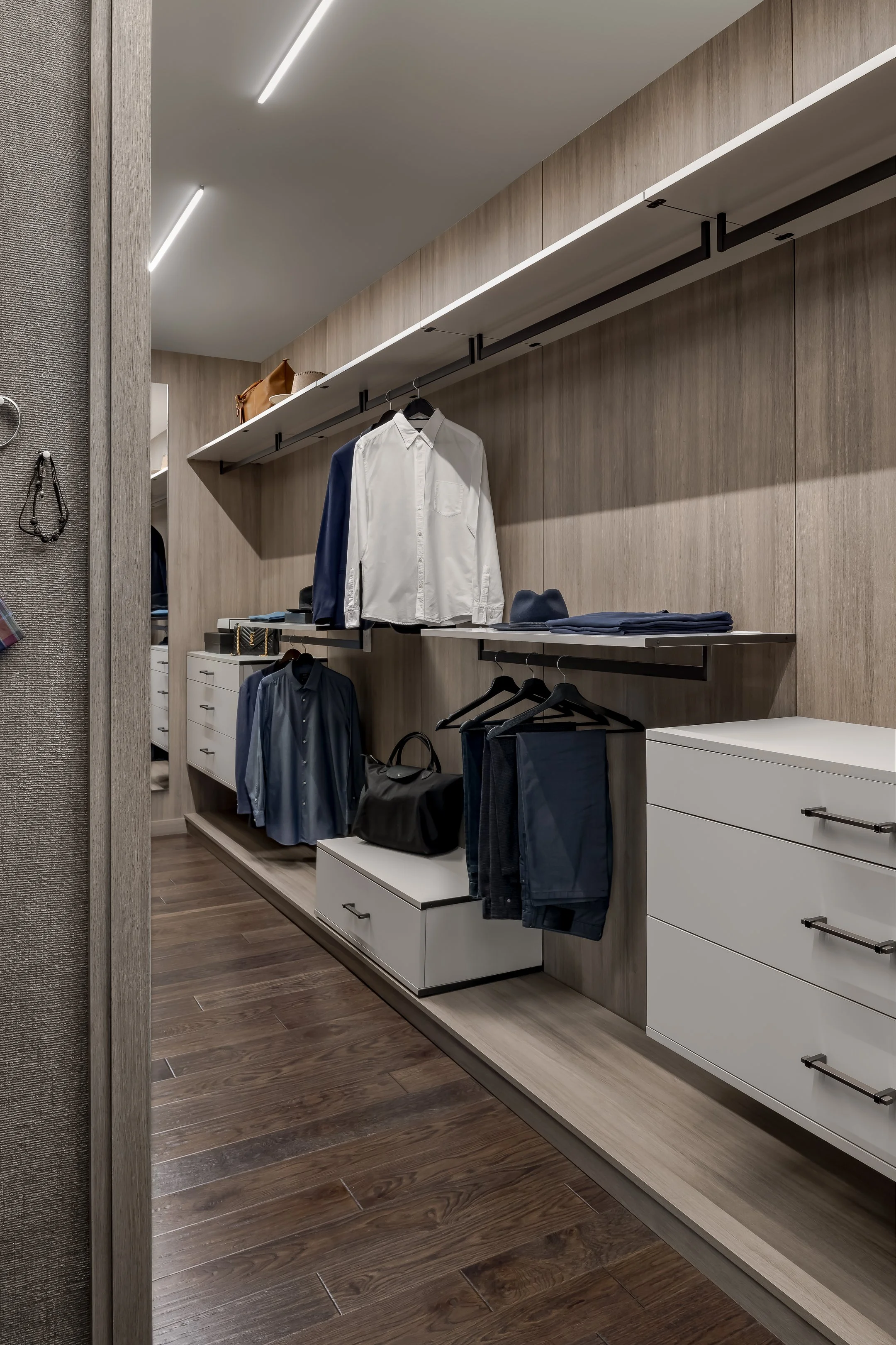Modern walk-in closet with wood-paneled walls, white shelves, hanging clothes, folded shirts, and hats, illuminated by sleek ceiling lights and featuring dark hardwood flooring.