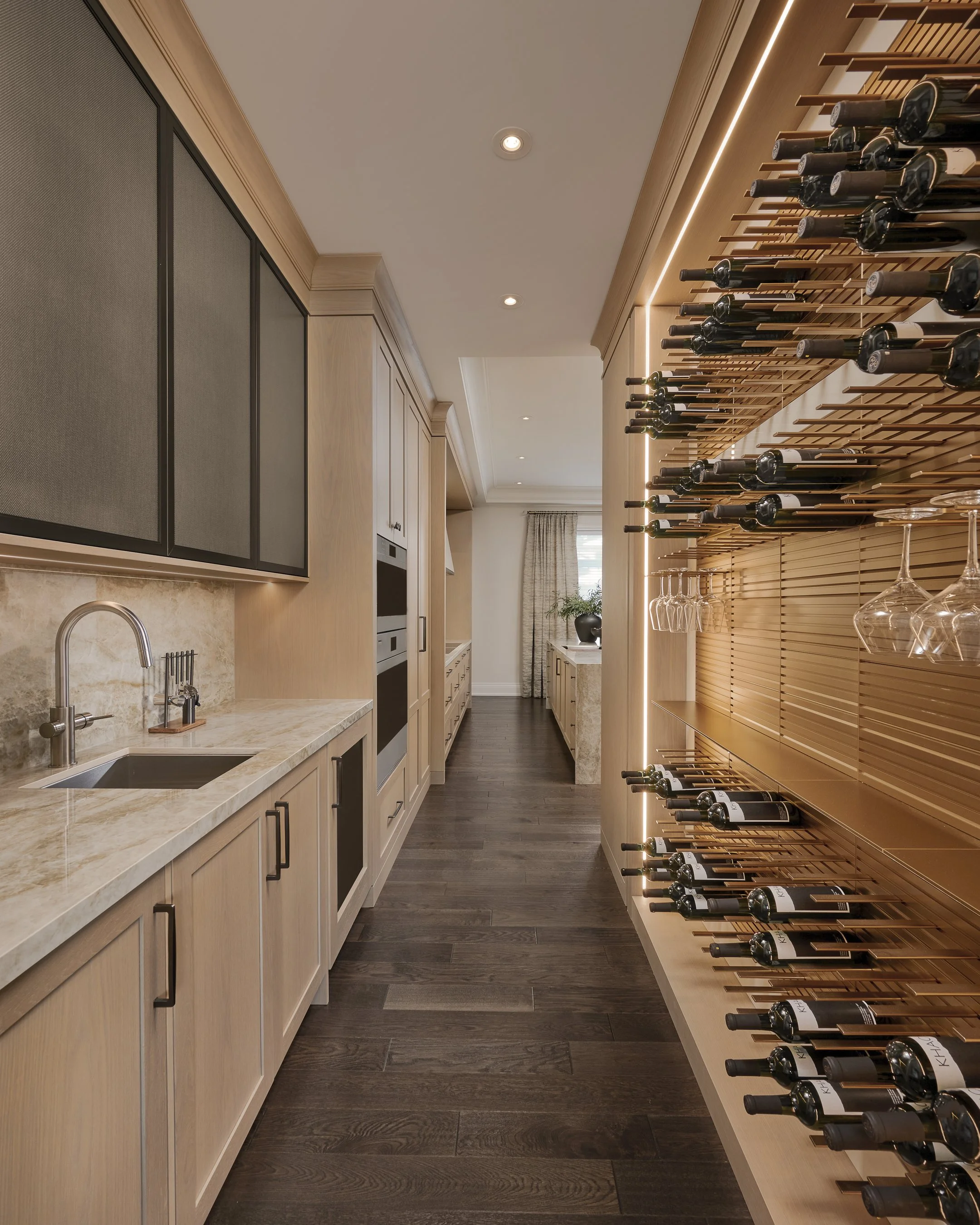 A modern wine cellar with a wooden wine rack on the right filled with wine bottles. The left side has a beige color kitchen cabinet with a marble countertop and a sink. The room has white walls, a white ceiling with recessed lighting, and a window with curtains at the end of the room.
