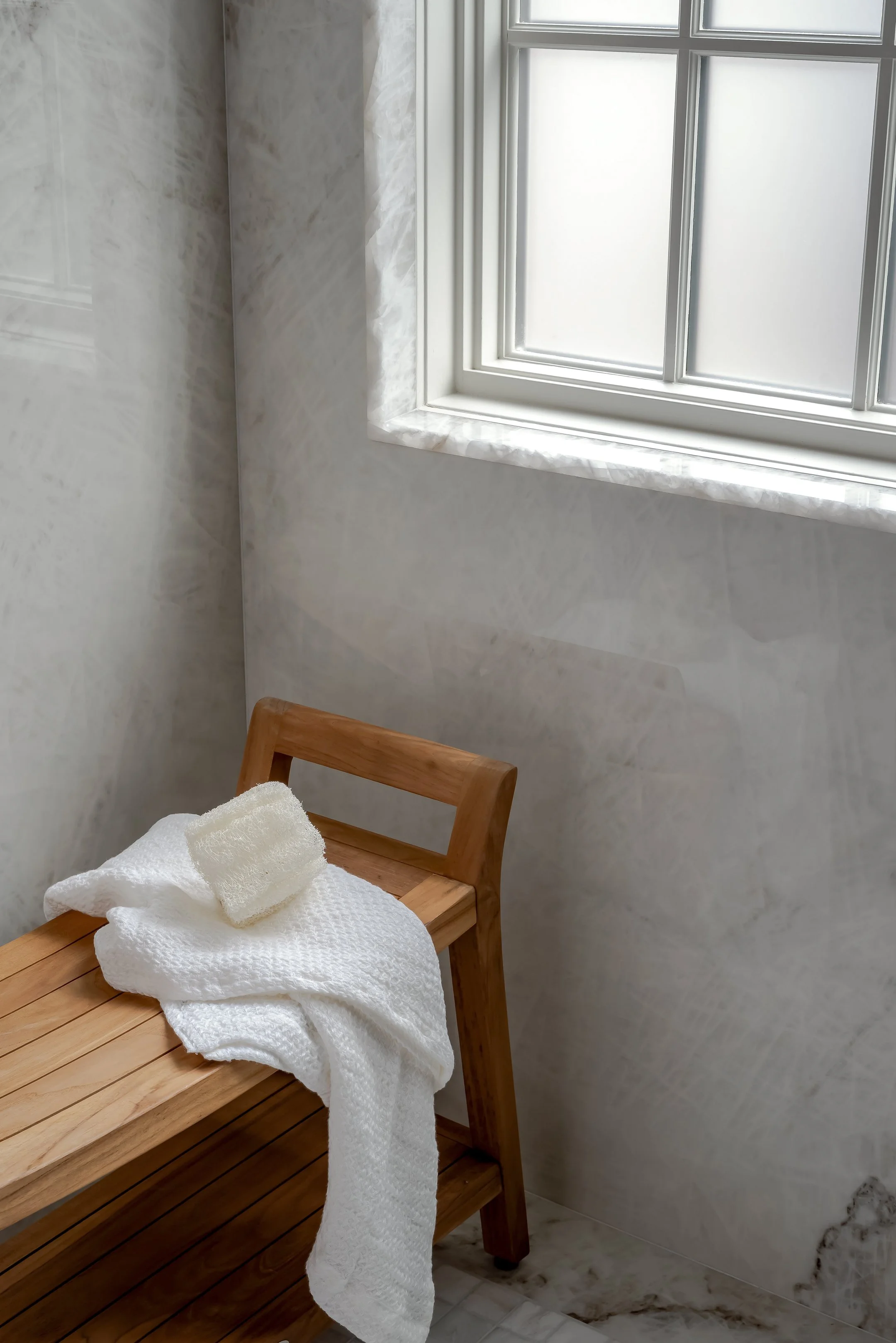 A wooden bench with white towels draped over it, in a room with light-colored walls, a window with frosted glass, and marble flooring.