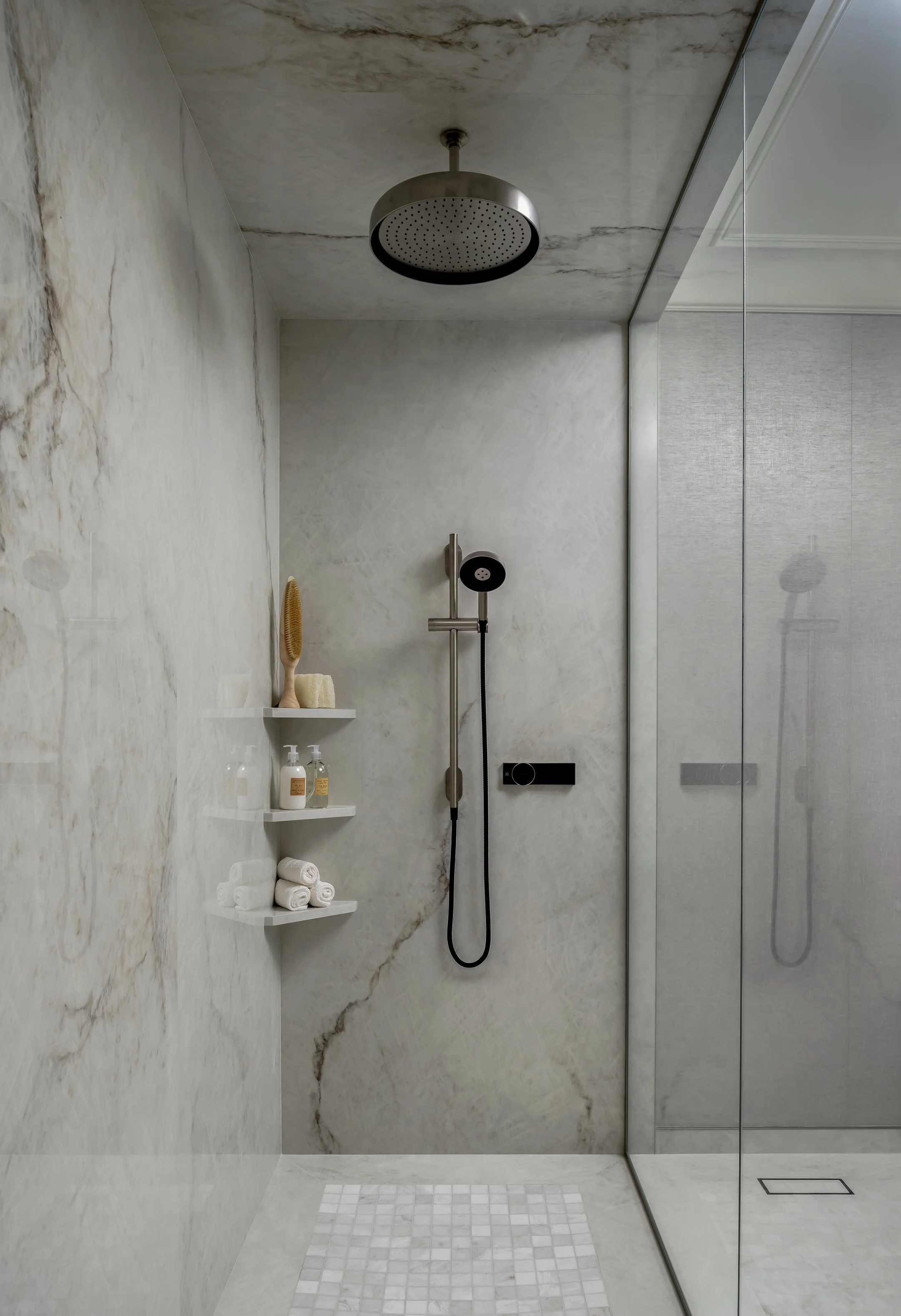 Modern walk-in shower with marble walls and floor, a rain shower head from the ceiling, and a handheld shower head. There are shelves with rolled towels and toiletries, and a glass door with a metal frame.