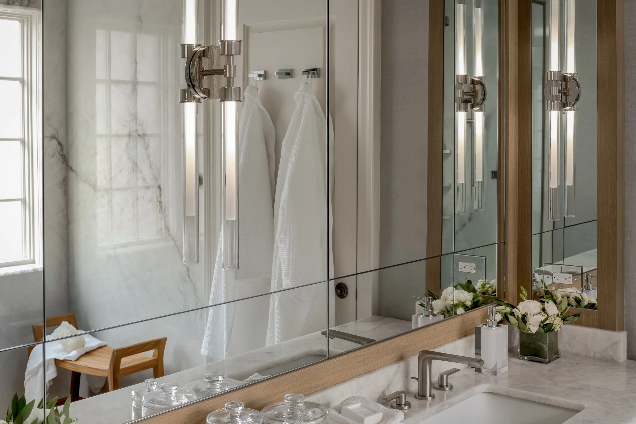 Bathroom mirror with modern light fixtures, flowers, and a view of a bathrobe hanging on a hook.