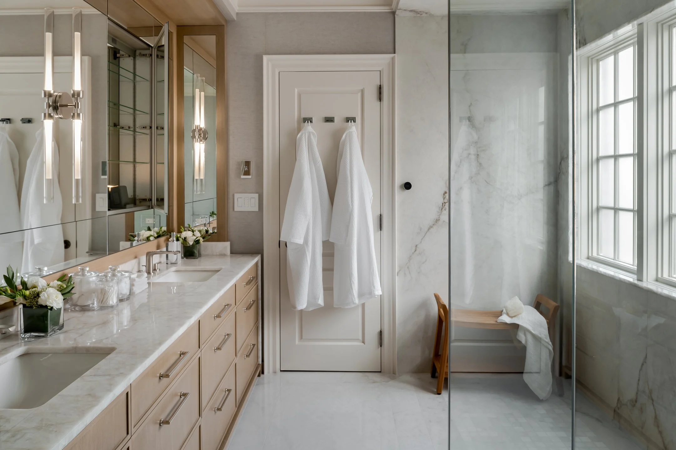 Elegant bathroom with a marble countertop, a large mirror, and a wall of glass shower with marble walls. Two white robes hang on hooks on a door, with a chair and a towel on it nearby. Natural light filters in through a window.