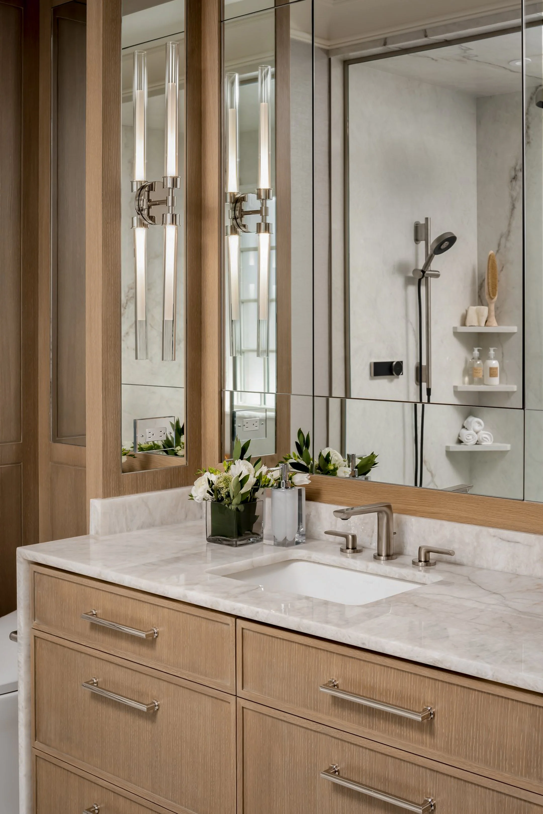 Bathroom vanity with a white marble countertop, a sink, a silver faucet, a vase with white flowers, and a bathroom mirror with modern light fixtures. The reflection shows a shower area with toiletries on shelves.