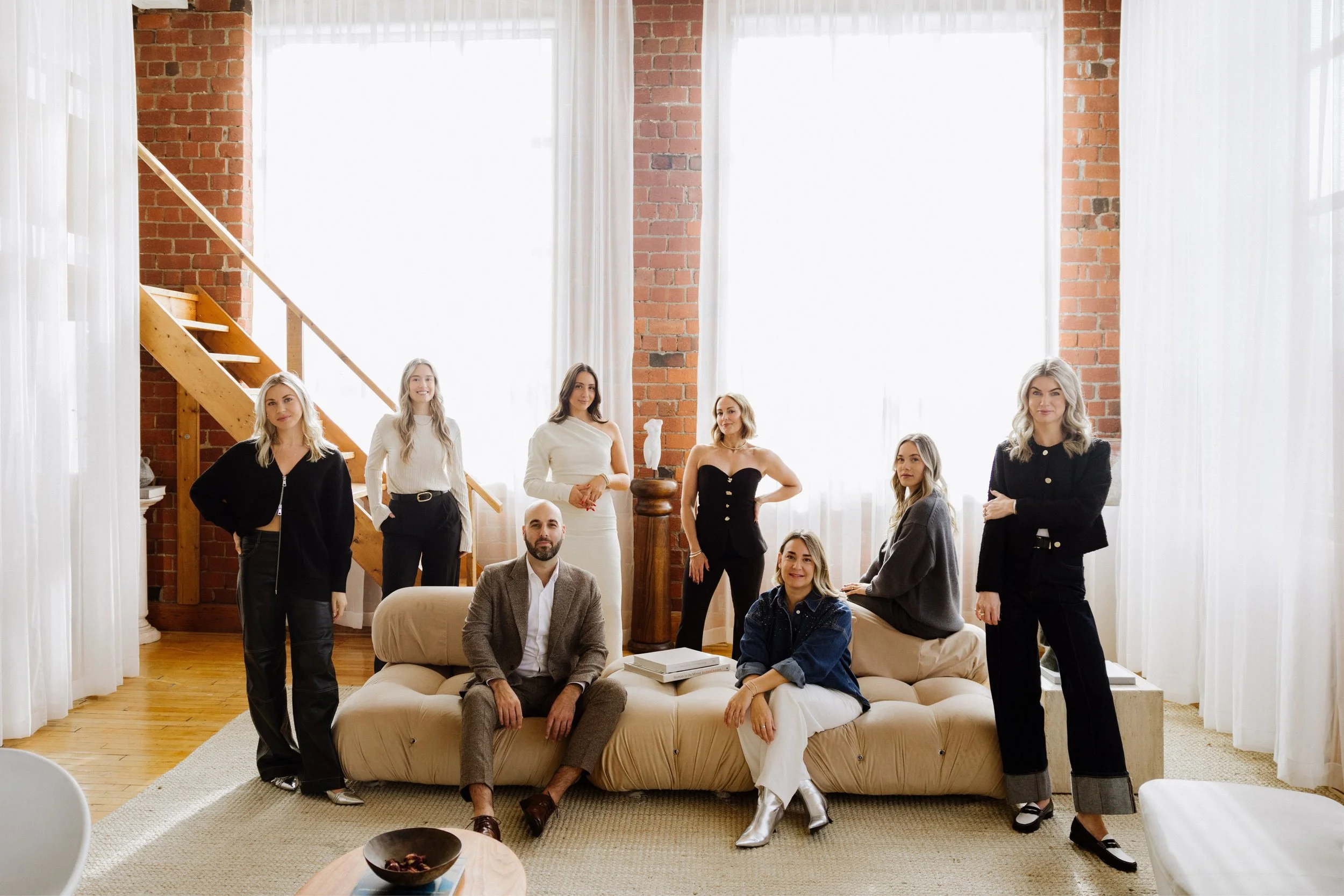 Group of nine diverse people in a modern living room with large windows, brick walls, and wooden staircase. They are posing around a beige sofa, some sitting and some standing, dressed in fashionable casual and business attire.