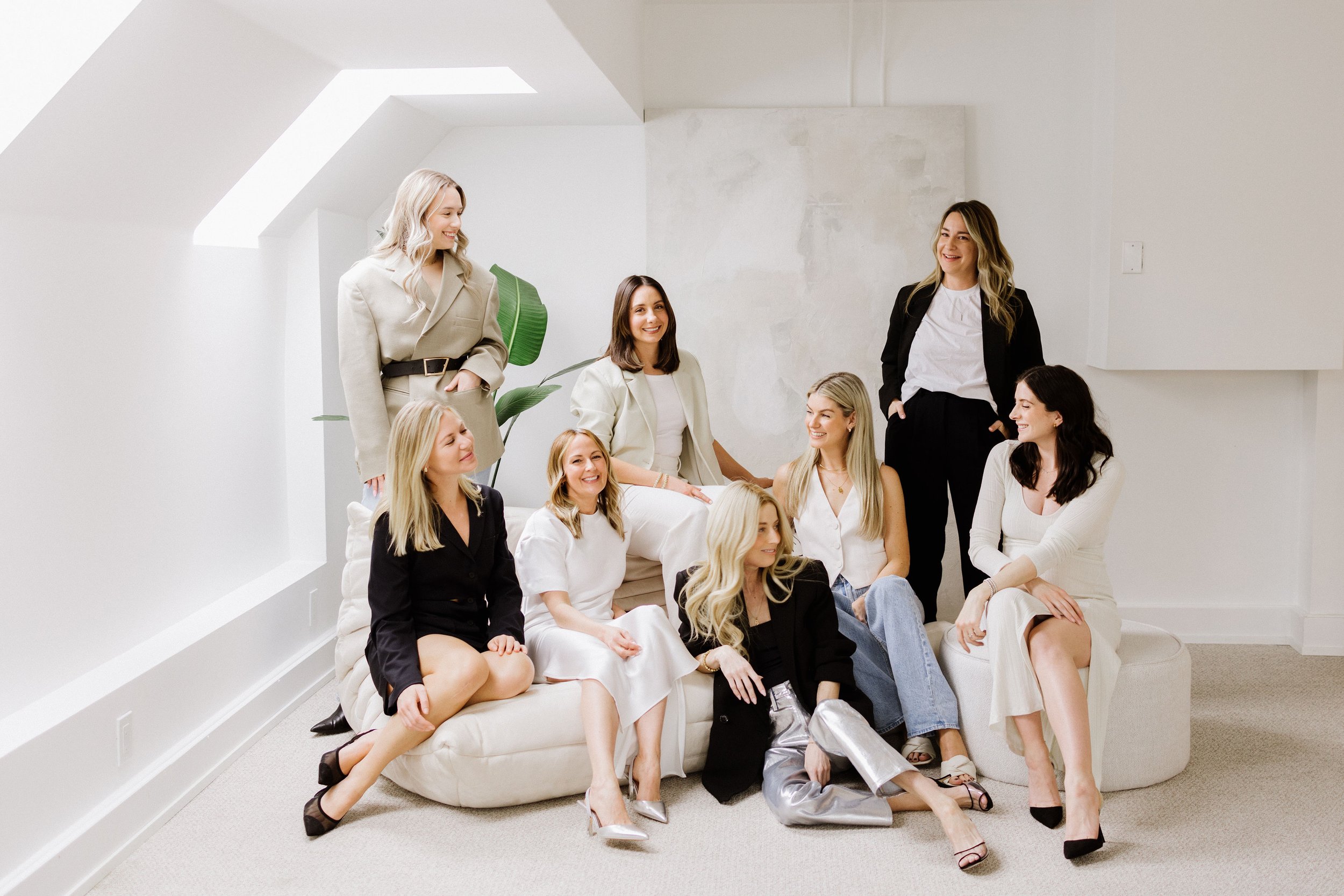 Nine women sitting and standing on a white sofa and in front of a plain white wall, engaging in conversation in a well-lit room.