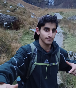 Young man outdoors in a natural setting with rocks and grassy terrain, wearing a black hoodie and carrying a backpack.