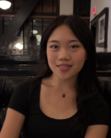 A young woman with long dark hair and a black top, smiling indoors.