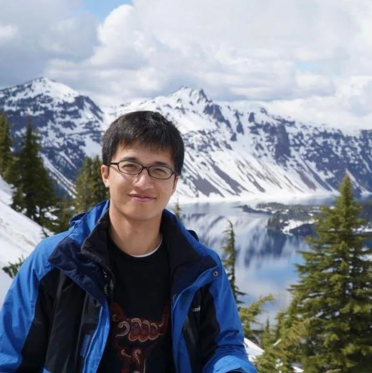 Person in a blue jacket standing in front of snow-covered mountains and a lake, with evergreen trees and cloudy sky in the background.