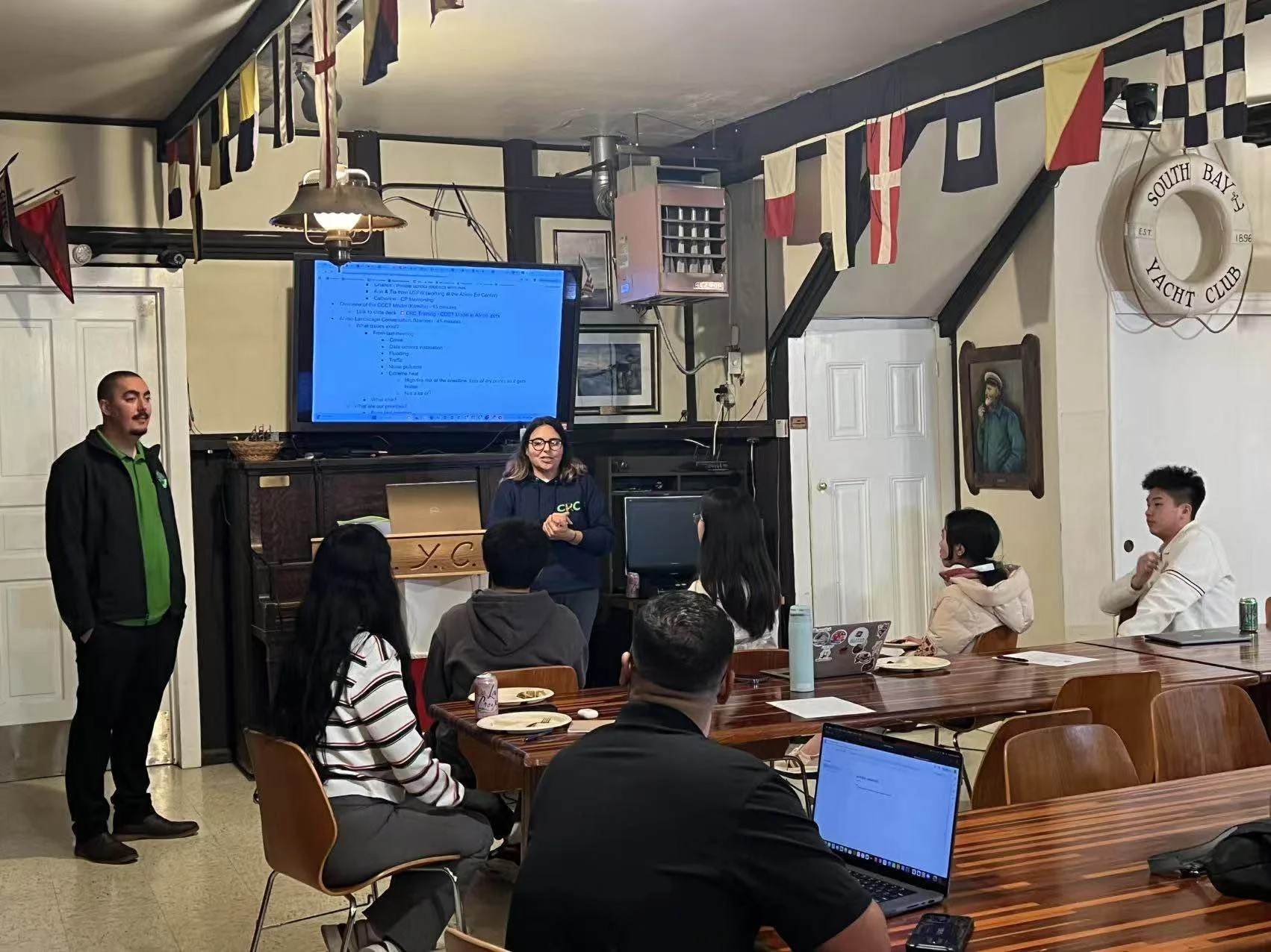 A group meeting in a room at the South Bay Yacht Club. A woman stands in front of a large screen displaying a presentation while others sit around tables, listening. Nautical flags are hung across the ceiling, and a laptop is visible on the table.