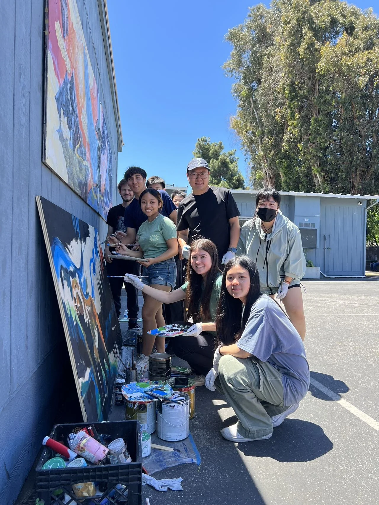 A group of people painting large colorful murals on an outdoor blue wall on a sunny day.