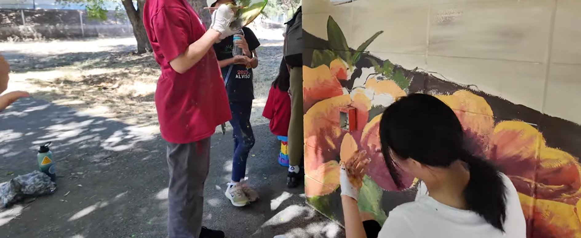 People painting a mural on a wall outdoors, with one person using a brush and others observing. Art supplies are visible on the ground.