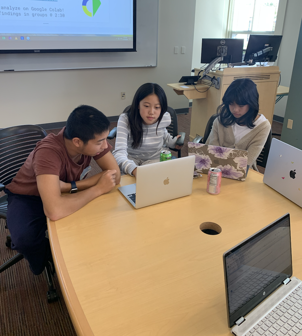 Three students sitting at a table in a classroom, working on open laptops. A projector screen displays a presentation in the background. The table has drinks and another closed laptop.