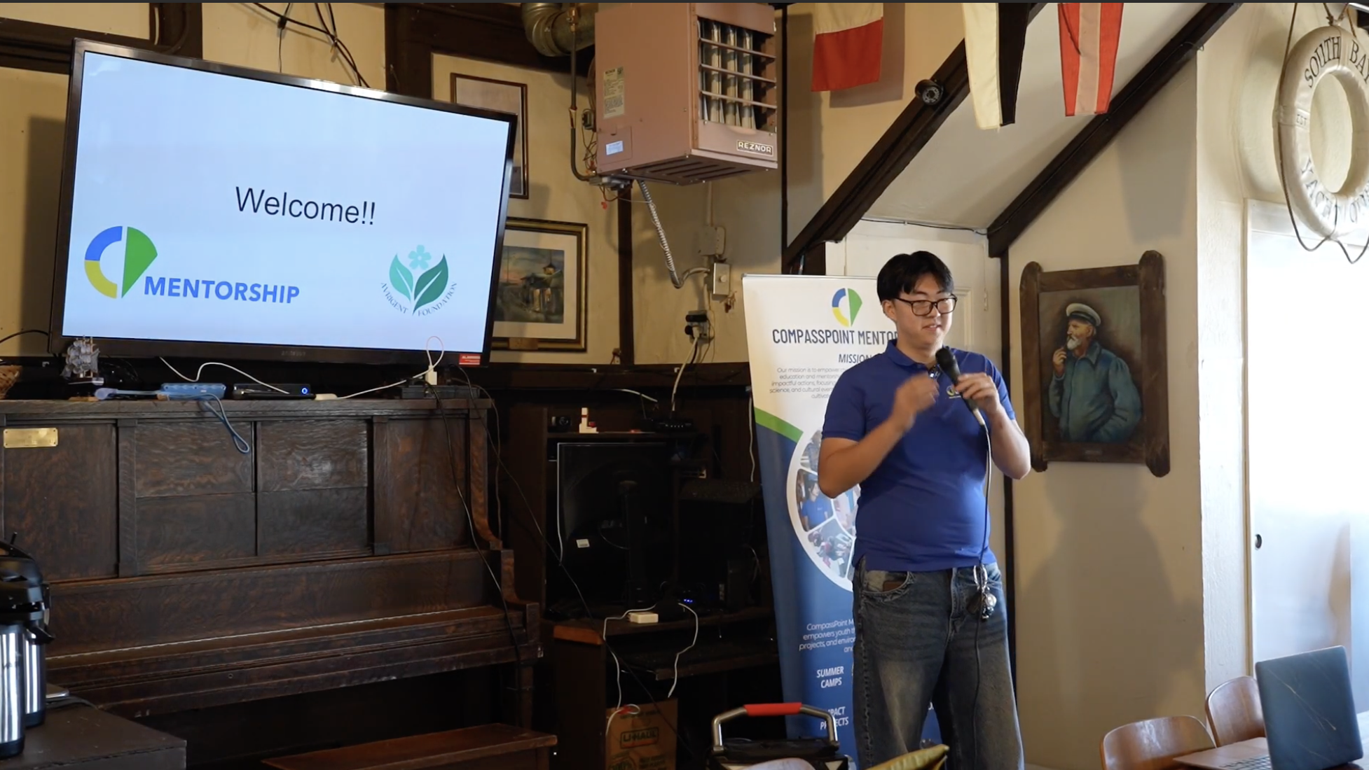 Person giving a presentation with a microphone in a room decorated with flags, a painting, and a piano. A TV screen displays 'Welcome!!' with logos for 'Mentorship' and 'Aubert Foundation'. A background banner reads 'Compasspoint Mentorship'."