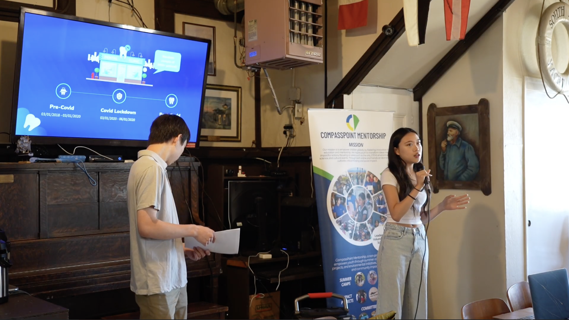 A young woman is speaking into a microphone during a presentation. A man is standing next to her looking at some papers. Behind them is a large screen displaying a timeline graphic with the phases "Pre-Covid," "Covid Lockdown," and "Post-Covid." Next to the screen is a banner for Compasspoint Mentorship. The room has wood paneling and nautical decor.