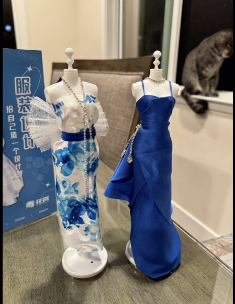 Two mannequin busts on a table displaying miniature dresses; one dress is white with blue floral patterns and a tulle embellishment, and the other is solid blue with spaghetti straps. A cat sits on a window sill in the background.