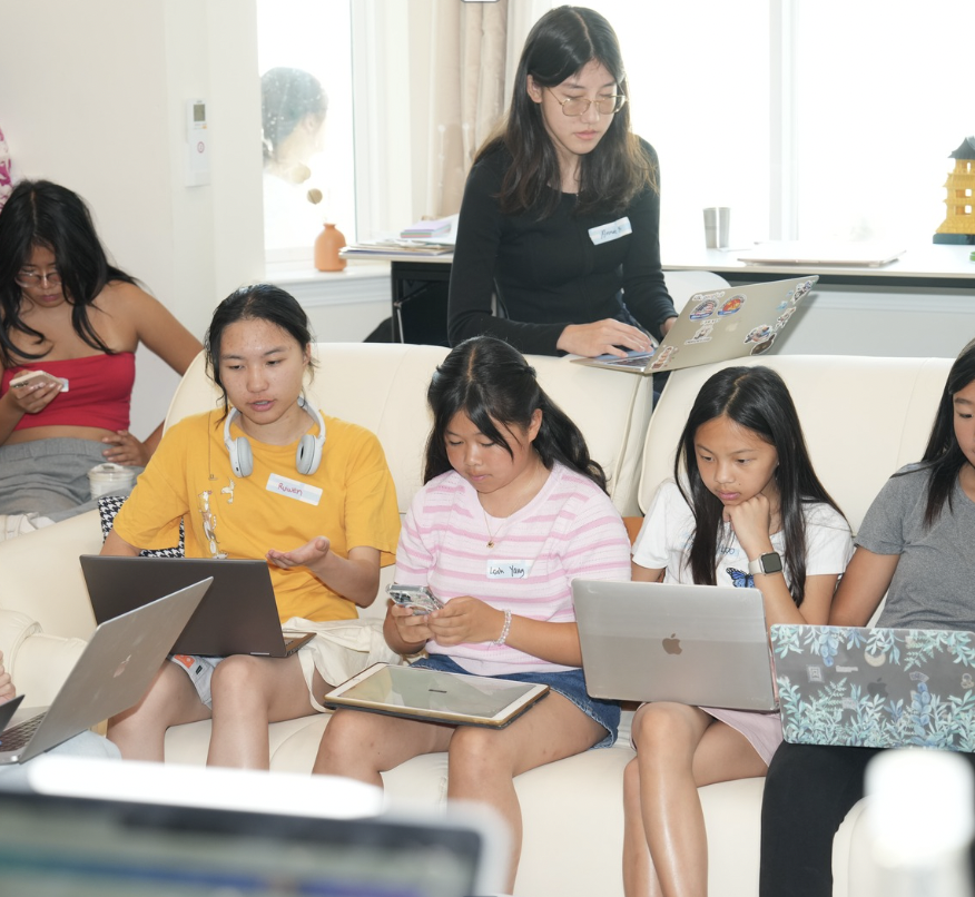 Group of young people sitting and standing with laptops and phones, engaged in a discussion or study session indoors.