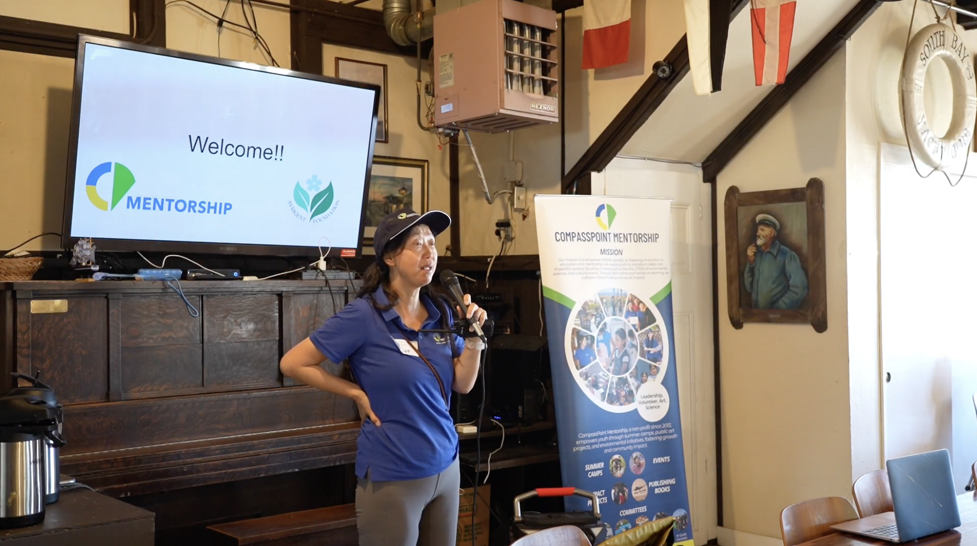 Woman in a hat speaking into a microphone at an indoor event. A 'Welcome' screen and a banner for 'COMPASSPOINT MENTORSHIP' are visible, along with a piano and nautical decorations. The setting appears casual and educational.