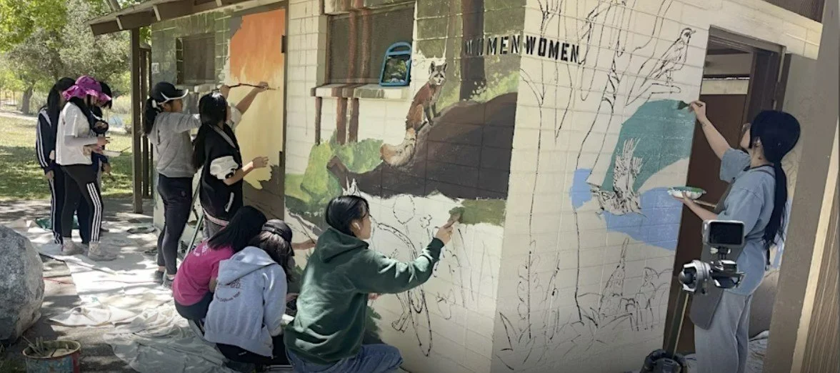 Group of artists painting a mural of animals and nature on an outdoor wall, some sketching and some adding color.