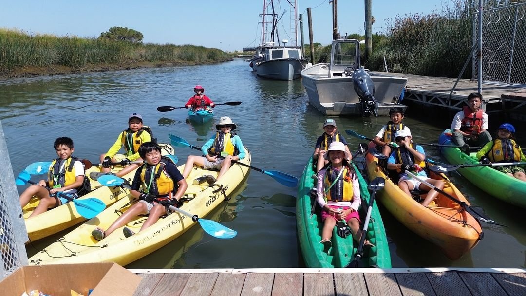 Group of children kayaking on a calm canal, wearing life vests and hats, with adult supervision in the background.