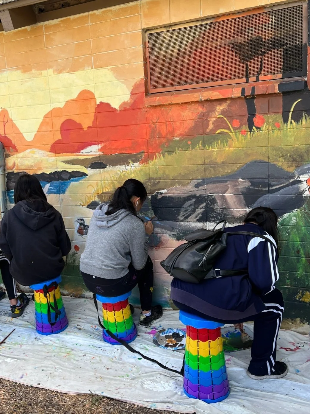 Three people are painting a mural on a brick wall, sitting on colorful, rainbow-striped stools, with painting supplies beneath them.