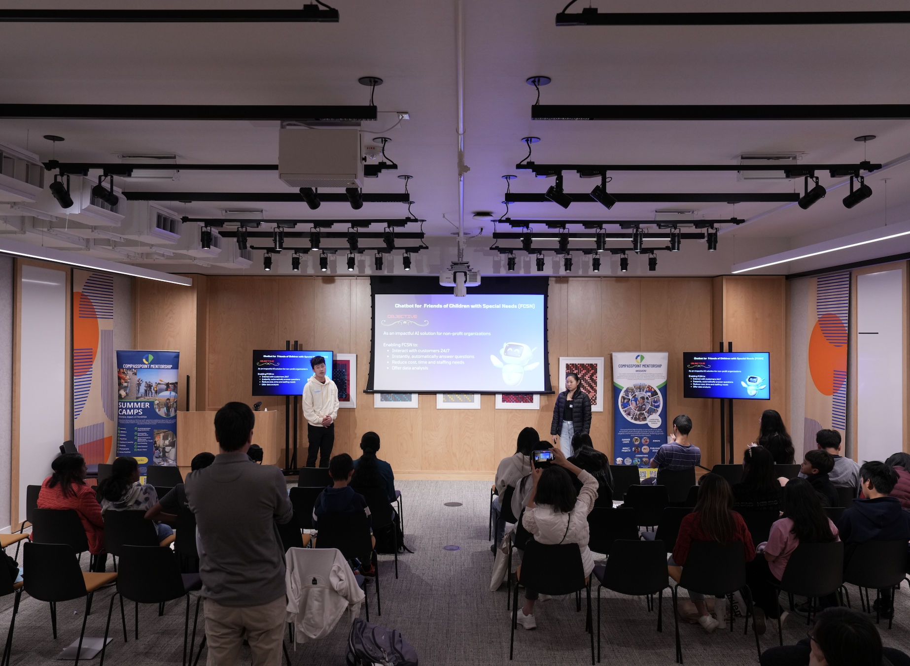 Group presentation in a modern conference room with audience. Two speakers are presenting slides on a screen titled "Chatbot for Friends of Children with Special Needs (FCSN)". Banners related to programs and events are displayed on either side.