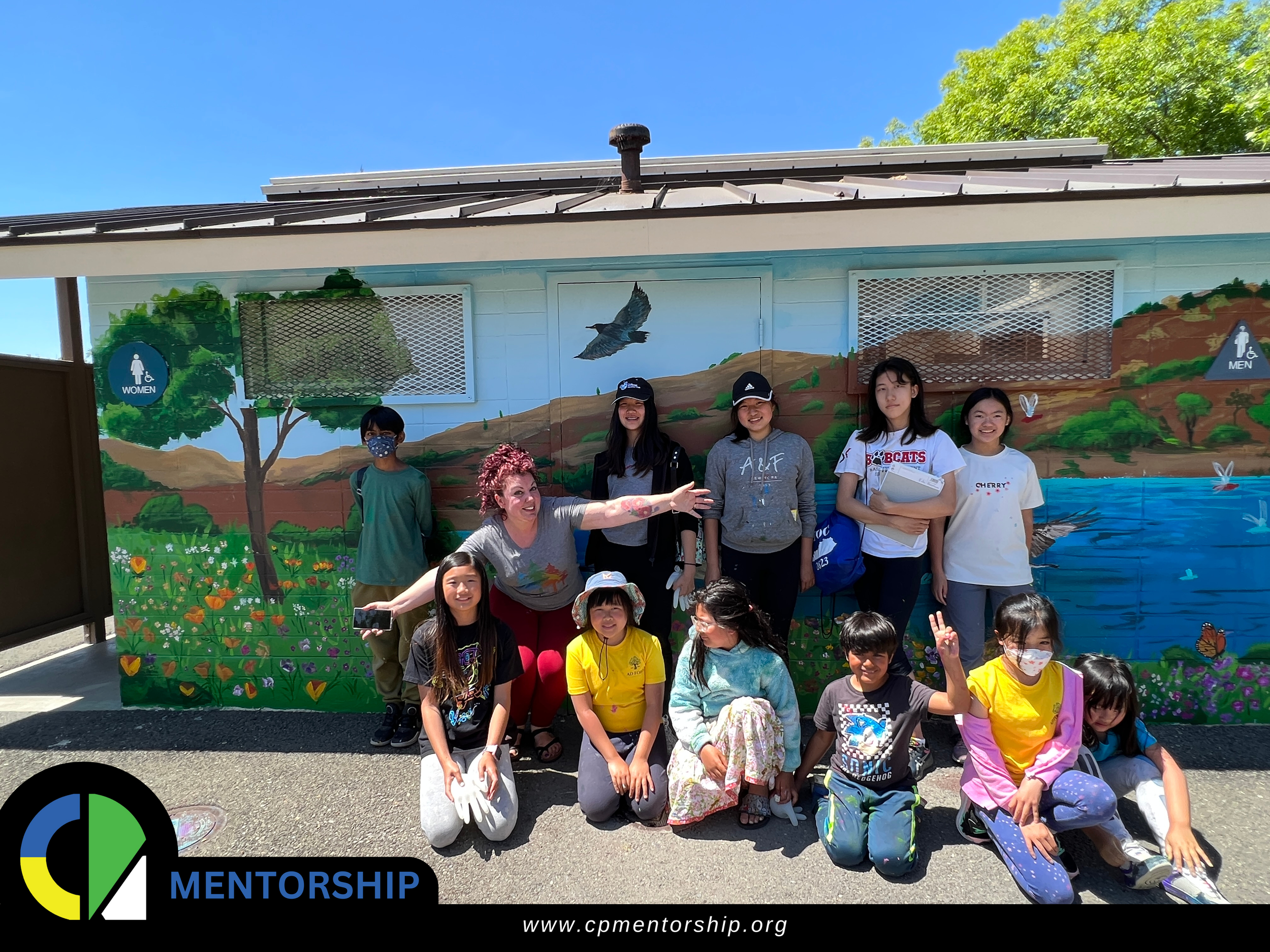 A group of people, including adults and children, posing in front of a building with a colorful mural depicting nature scenes. The mural features trees, a landscape, and a body of water. The individuals are smiling and appear to be part of a mentorin