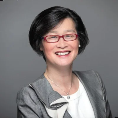 A woman with black hair, glasses, and a gray jacket smiling against a gray background.