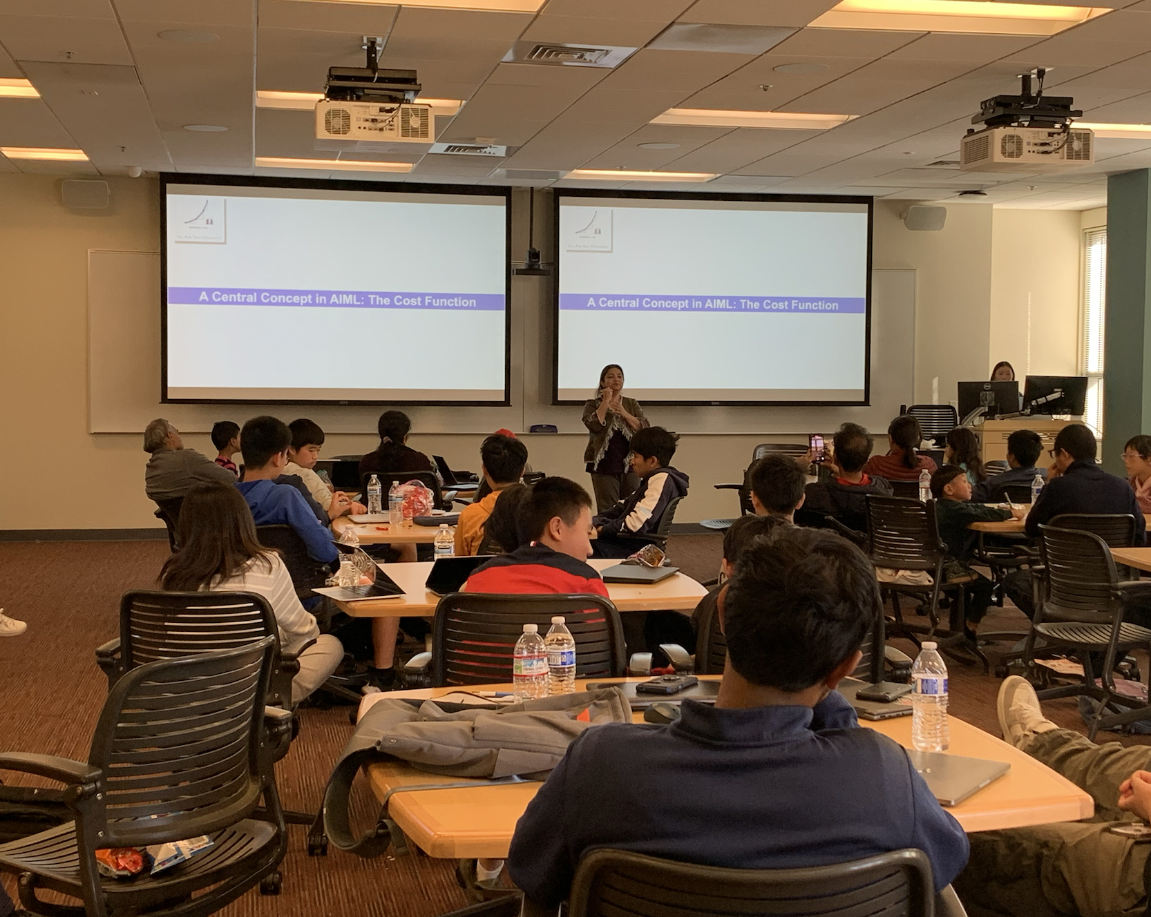 People attending a lecture in a classroom with a presentation on a topic related to AI/ML cost function. Several tables with students sitting, notebooks, and snack items are visible.
