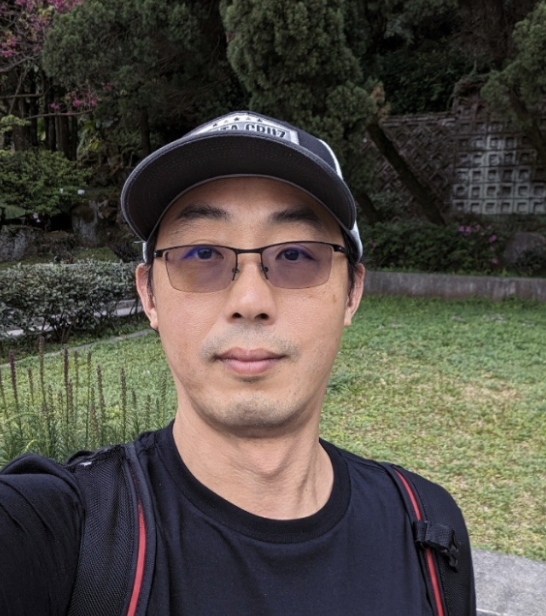 Man taking a selfie outdoors, wearing glasses, a baseball cap, a black shirt, and a backpack, with trees and greenery in the background.