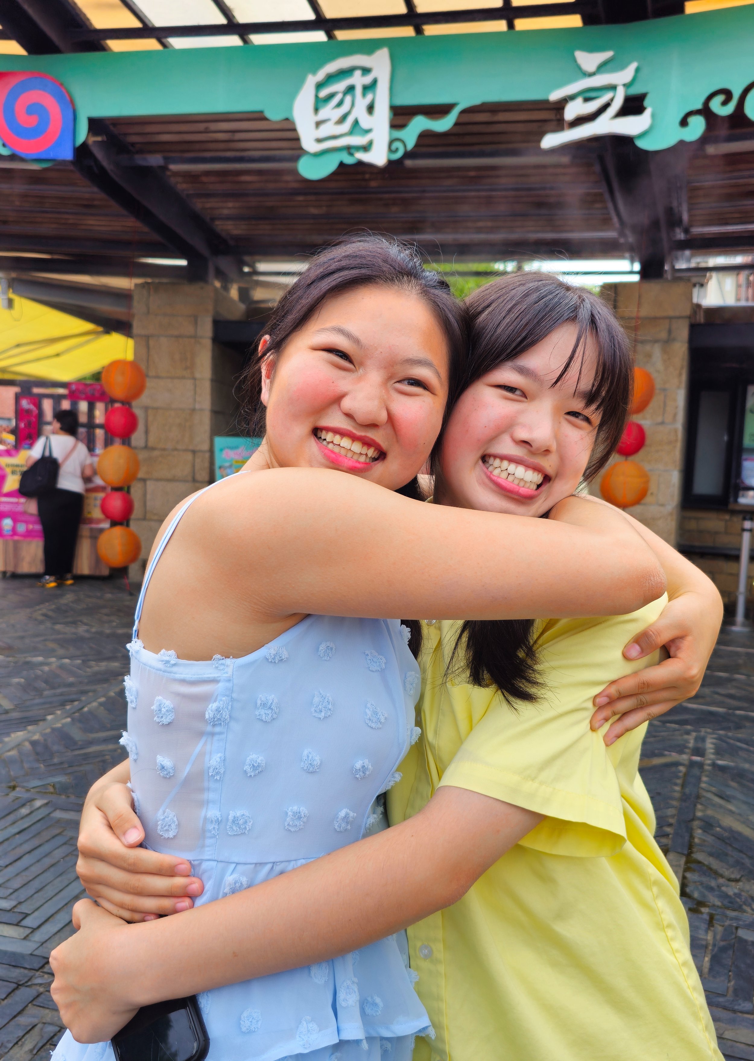 Two smiling friends hugging outdoors in front of a colorful sign.
