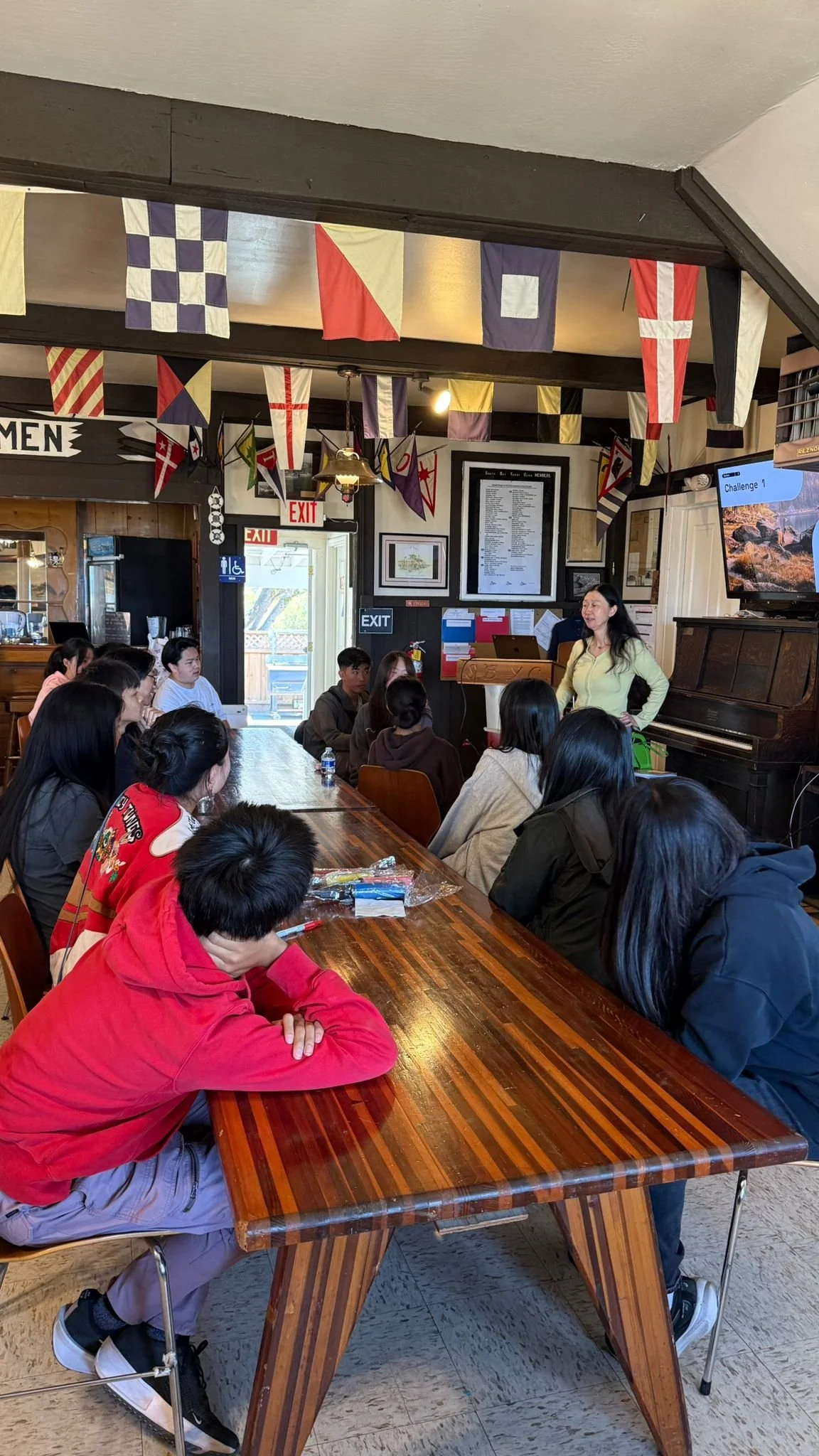 A group of people sitting around a wooden table in a restaurant or cafe, listening to a woman speaking at the front. The room is decorated with nautical flags hanging from the ceiling, and there is a piano against the wall. On the wall behind the speaker, there is a large framed menu, and a TV screen displays a challenge in an outdoor setting.
