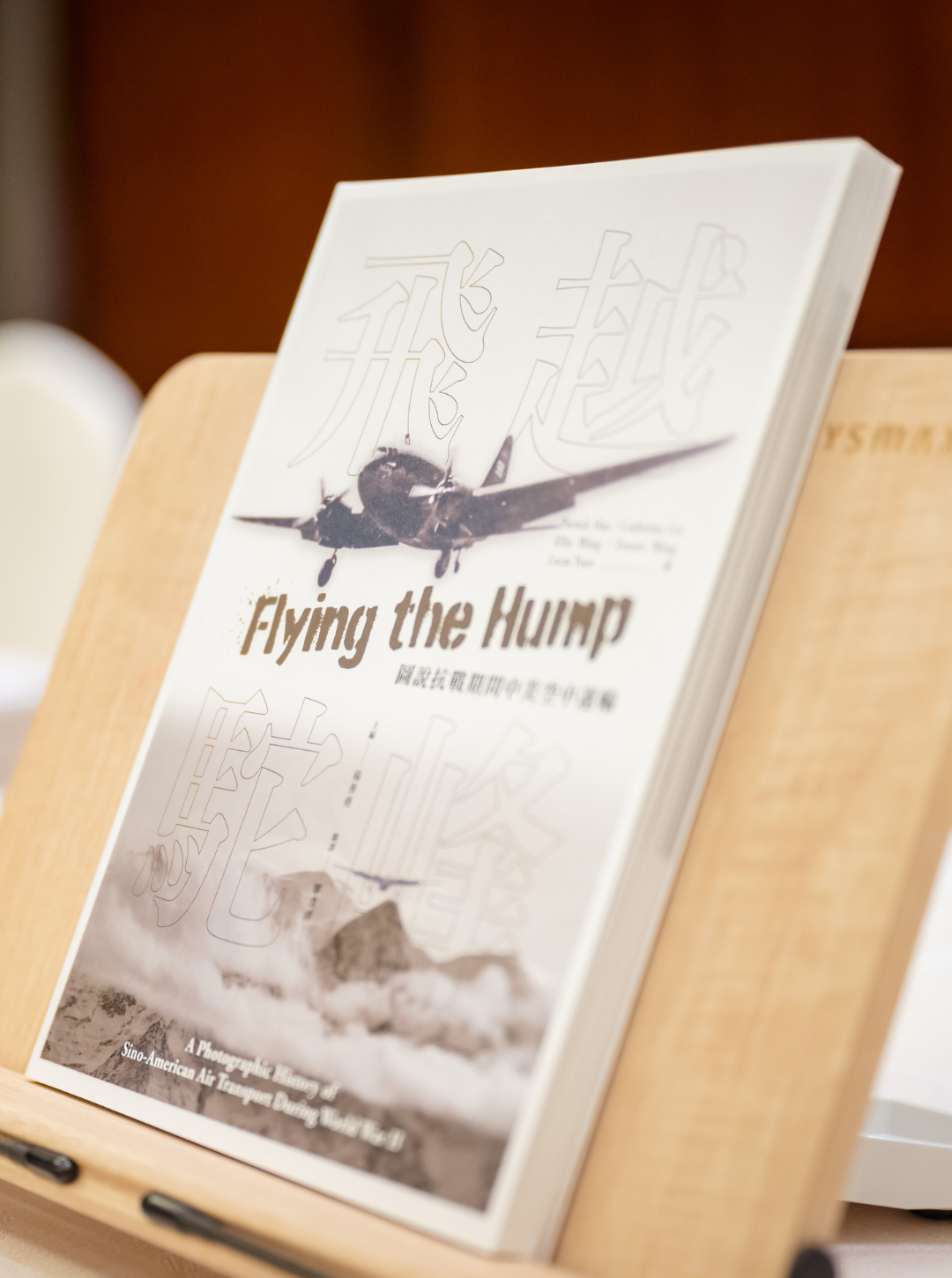 Book titled 'Flying the Hump' displayed on a wooden stand, containing black and white imagery of a plane and mountains, with English and Chinese text.