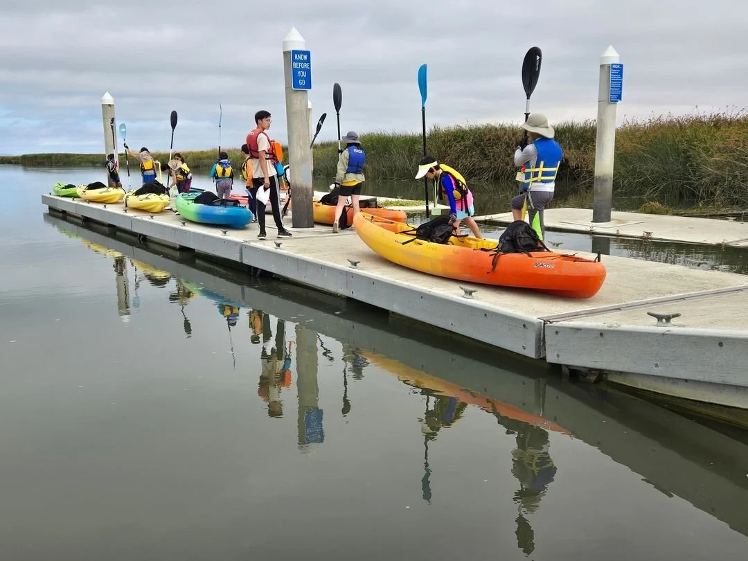 Group of people preparing kayaks on a dock in a calm waterway, wearing life jackets and holding paddles, with grassy banks in the background.
