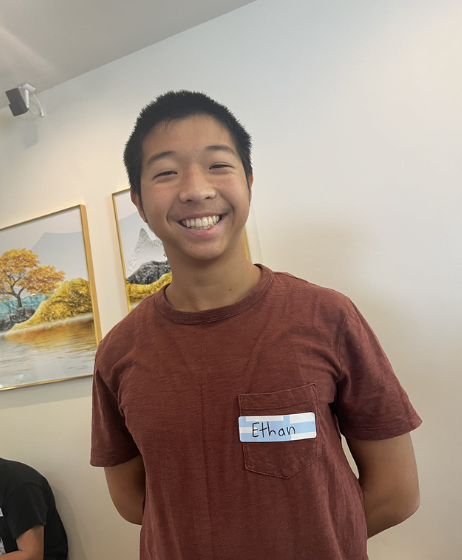 Smiling person in a red shirt with a name tag, standing indoors with framed art in the background.