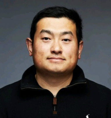 Portrait of a man with short black hair, wearing a black zip-up jacket, against a muted background.