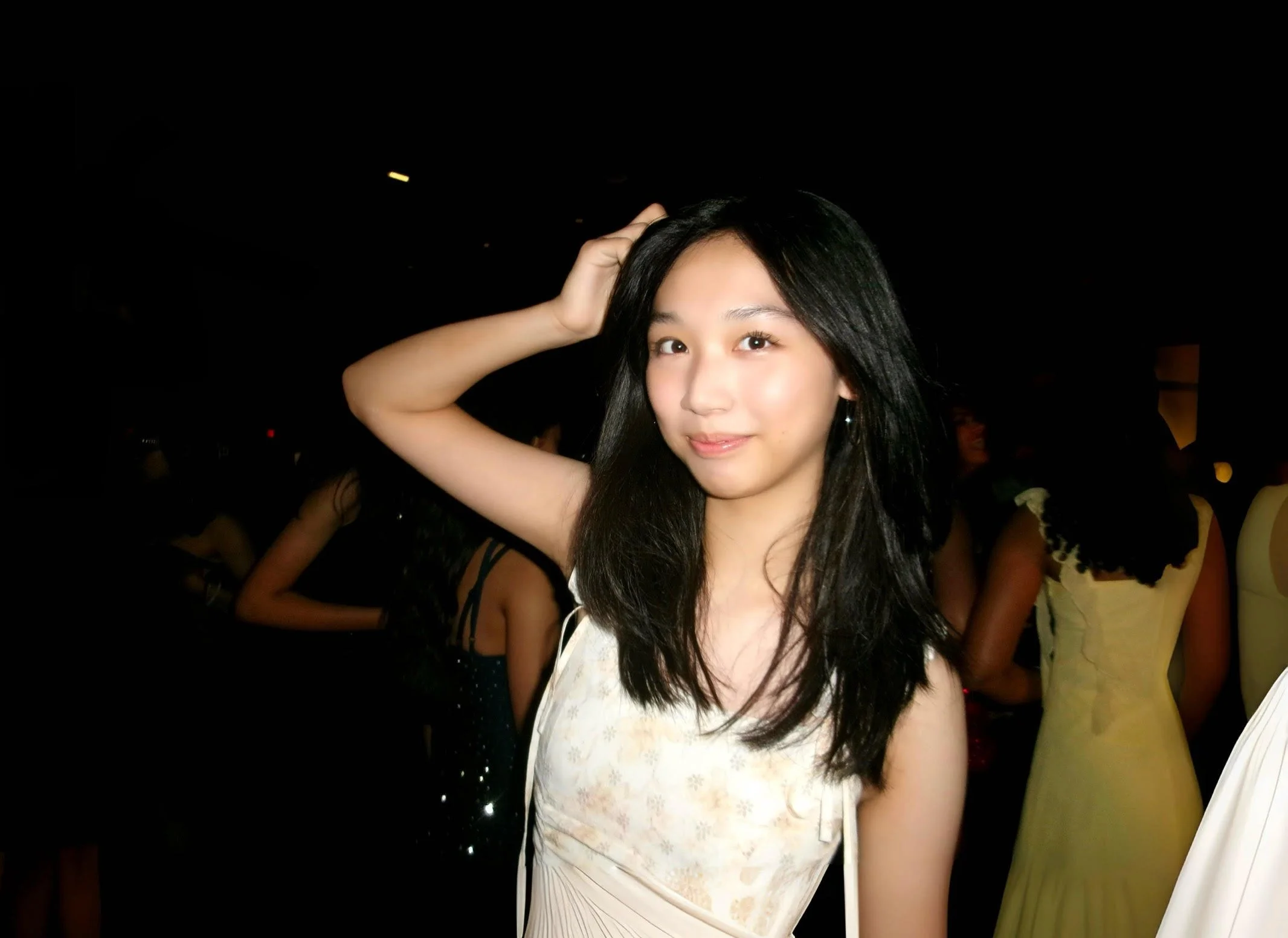Young woman with black hair in a sleeveless light-colored dress, standing in a dark room with other people in the background.