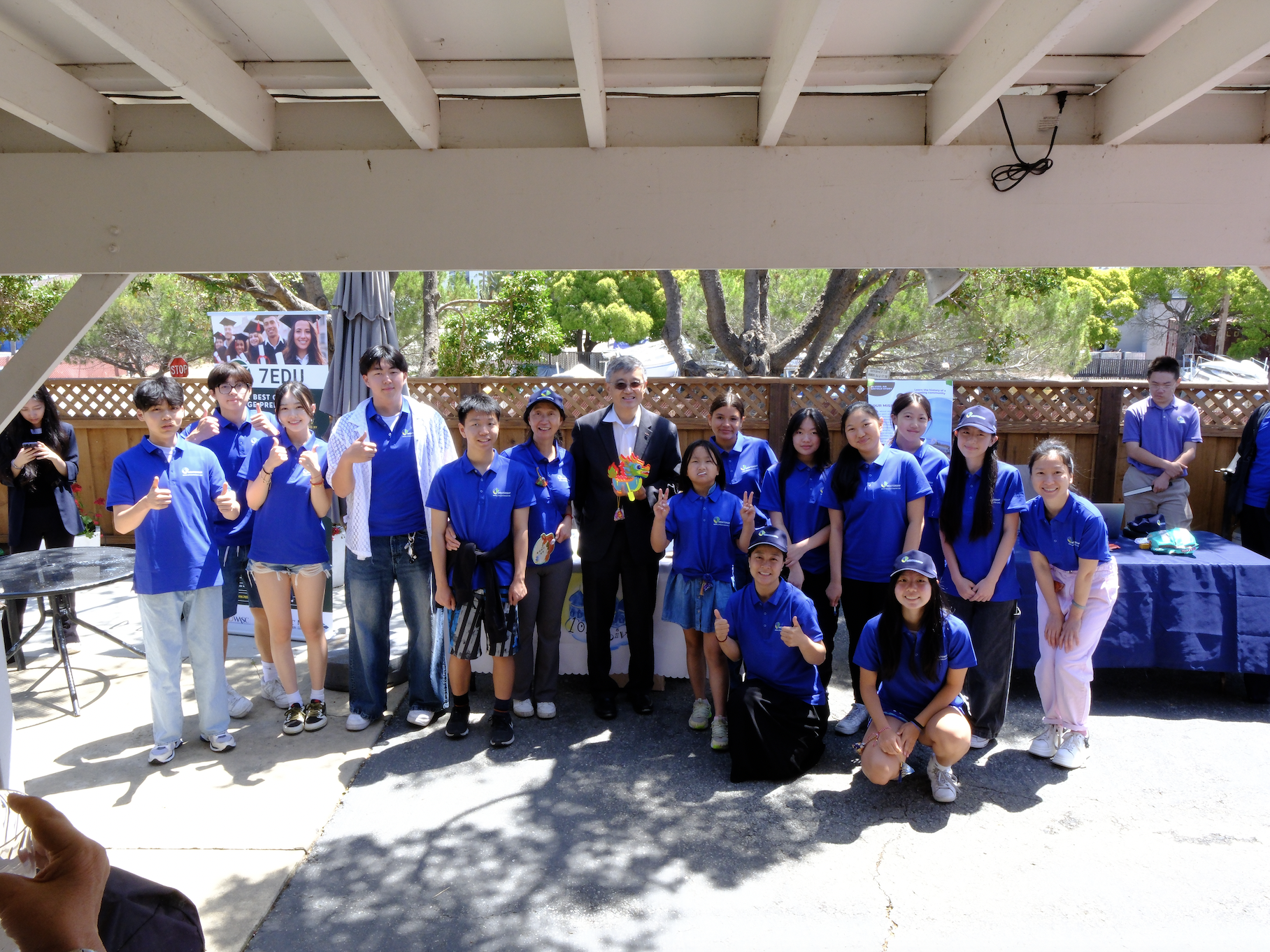 Group of people in matching blue shirts posing outdoors under a patio, with a wooden fence and trees in the background.