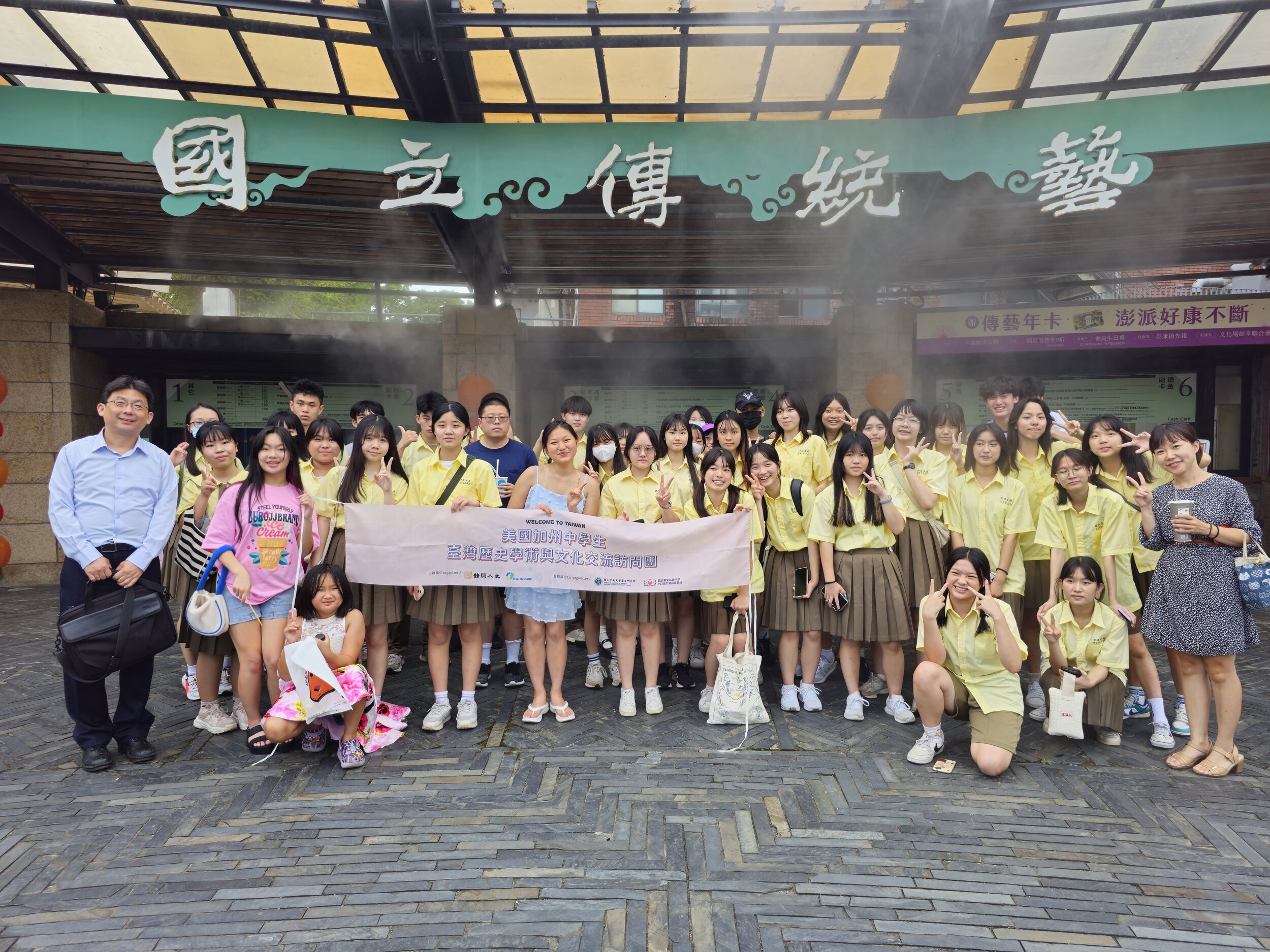 A group of students and adults, mostly wearing yellow uniforms and casual clothes, posing in front of a building with Chinese characters overhead. They hold a banner with writing in English and Chinese. The scene is informal and outdoors, with tiled